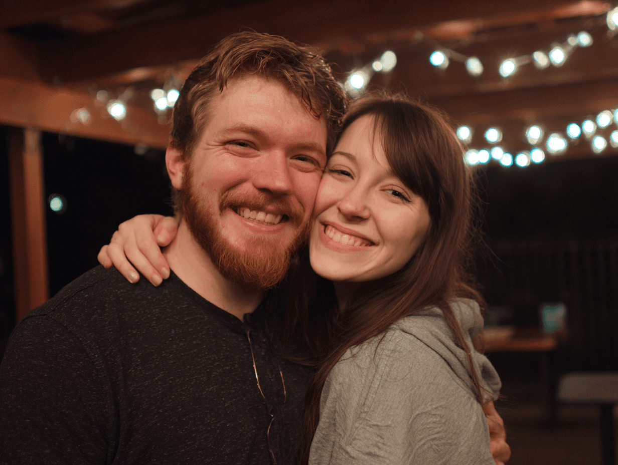 A smiling couple embraces in a warm, illuminated setting with string lights in the background.