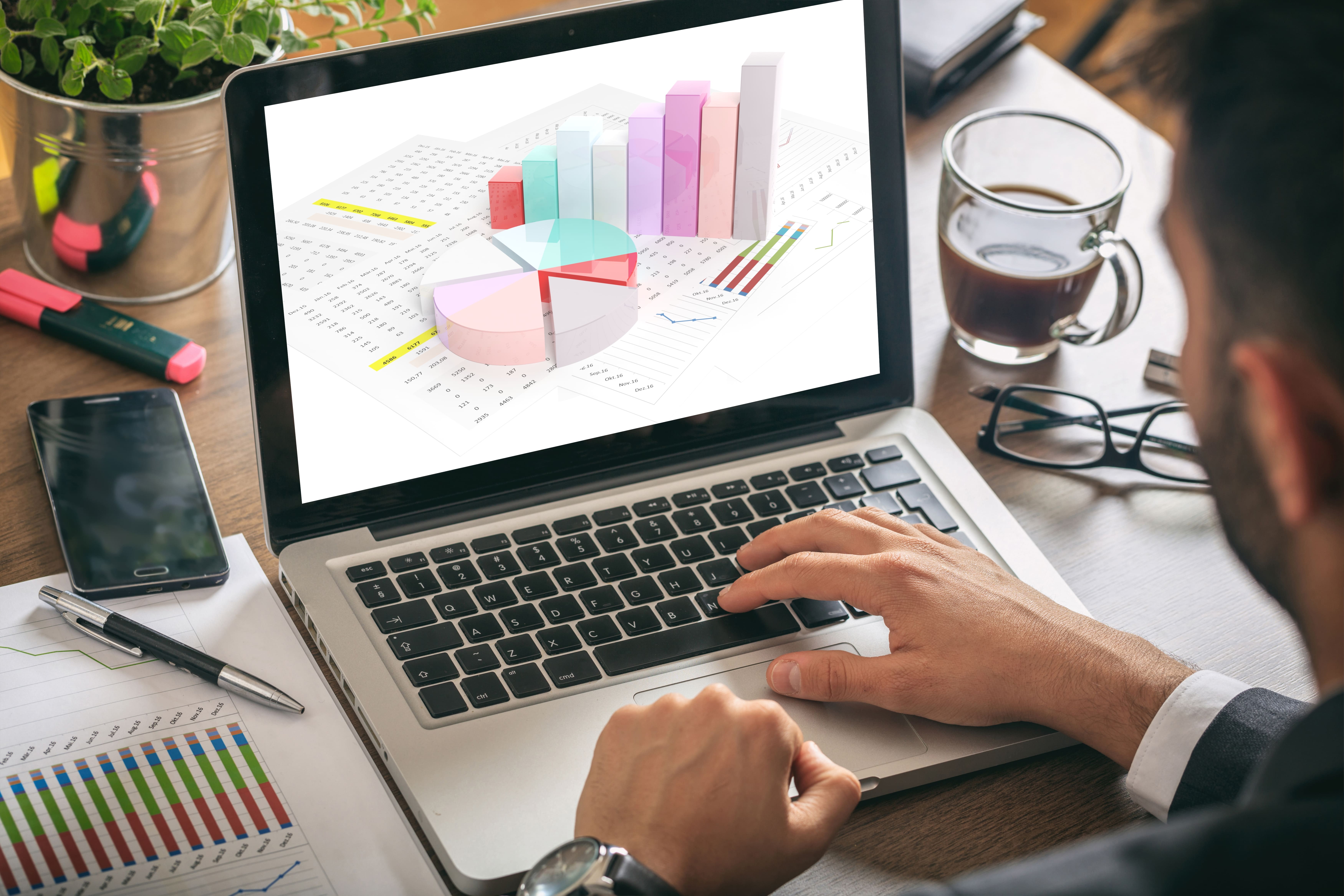 A imagem mostra uma pessoa usando um laptop em uma mesa de trabalho. Na tela do laptop, há gráficos em 3D, incluindo um gráfico de barras e um gráfico de pizza, com várias cores claras e pastéis. Ao lado do laptop, há uma folha de papel com gráficos impressos, um smartphone, uma caneta e marcadores em cores rosa e amarelo. Também há uma xícara de café meio cheia e óculos de leitura. O ambiente sugere um espaço de trabalho organizado, provavelmente relacionado à análise de dados ou finanças.