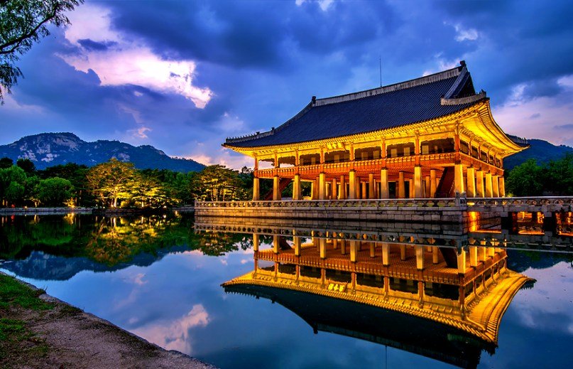 A wooden desk by a window, with sunlight illuminating a laptop, notebook, and a decorative vase.
