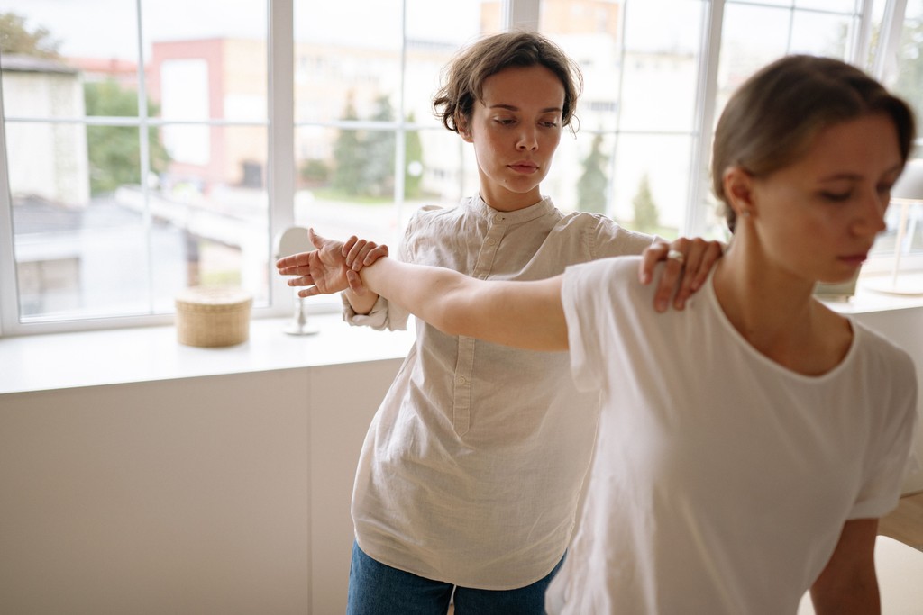 A physiotherapist guiding a client's arm and shoulder through a posture exercise beside a window.