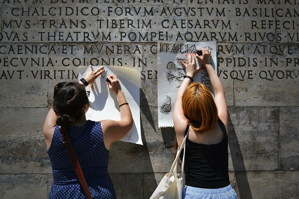 students shaving the graphite pen to a paper and put over an engraved text on the wall