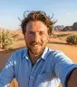 A high-angle selfie of a smiling man with light brown, windswept hair and a groomed beard, standing in a vast desert. He is wearing a light blue linen button-down shirt. The background features rolling golden sand dunes, sparse green desert shrubs, and rugged sandstone mountains under a clear, bright blue sky.