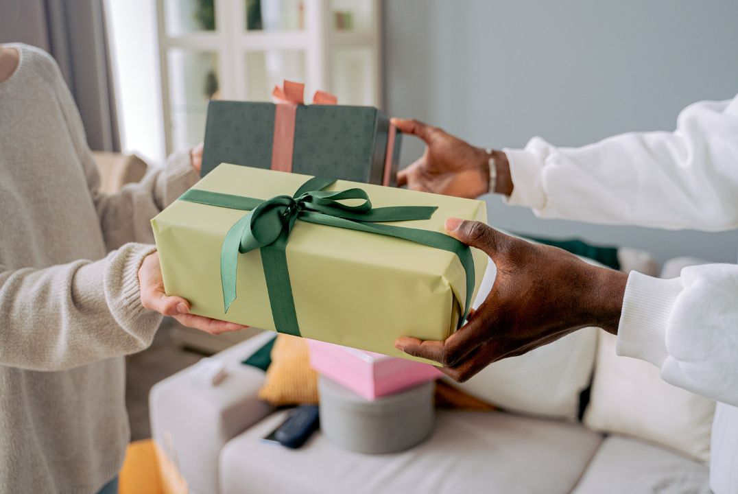 Two people exchanging a beautifully wrapped gift with a green ribbon, symbolizing appreciation and the joy of giving in a cozy, comfortable indoor setting.