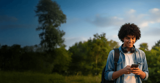 A teenage boy, standing in a park, smiling and looking in his phone.