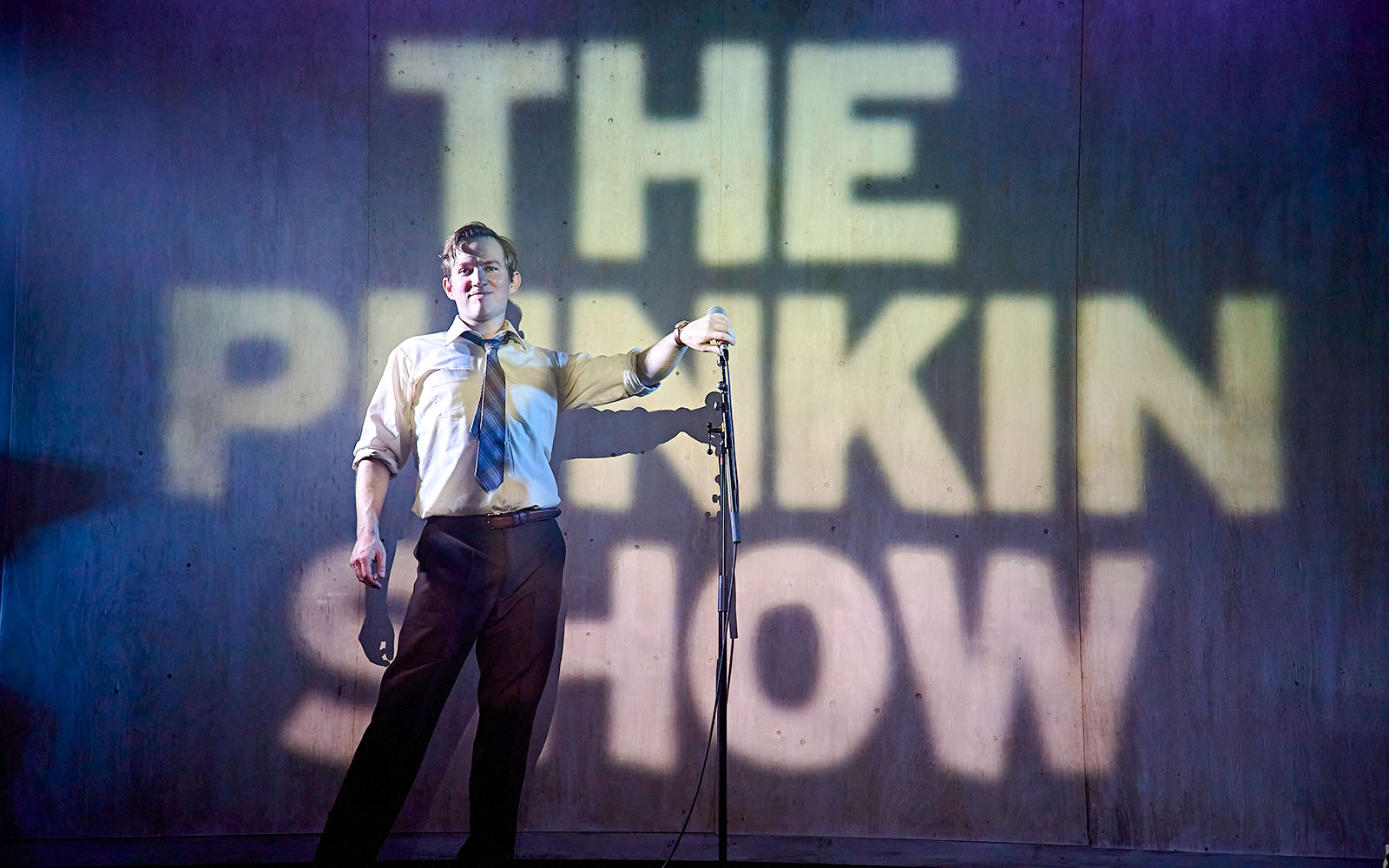 Performer on stage with "The Pinkin Show" projected behind, holding a microphone.