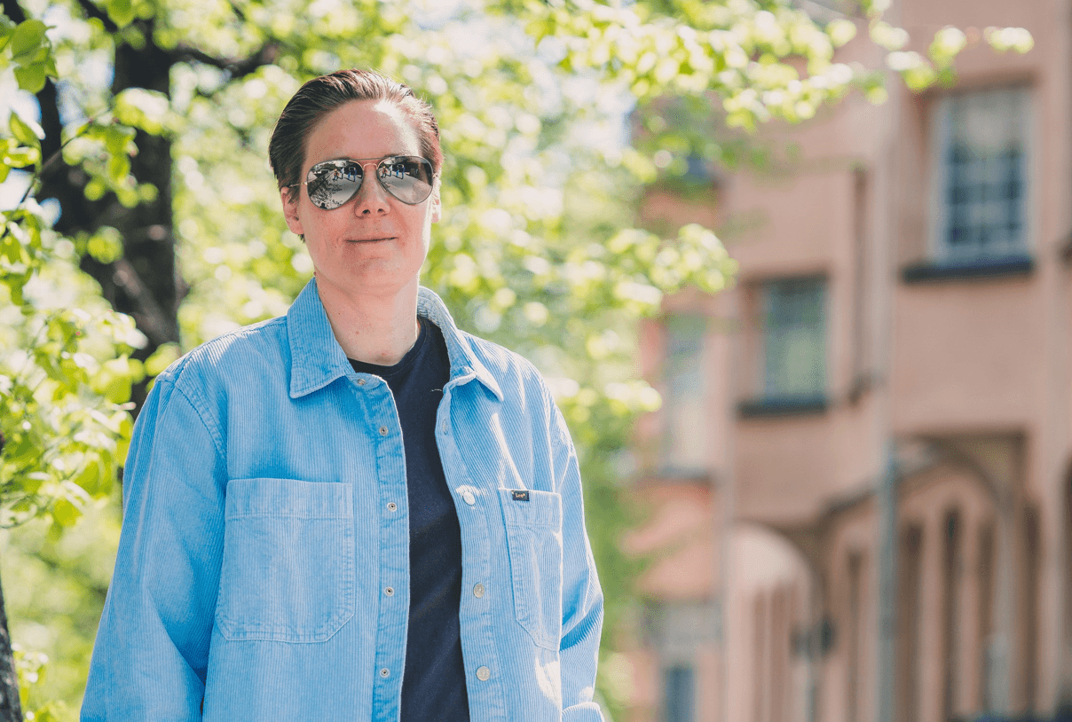 Tuulia Virhiä, CEO of SwanIT, wearing sunglasses and a light blue denim shirt, standing outdoors in front of some trees and a building