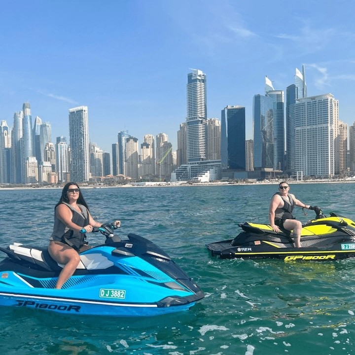 Two riders on jet skis near the Dubai Marina waterfront, with modern high-rise towers in the background on a sunny day