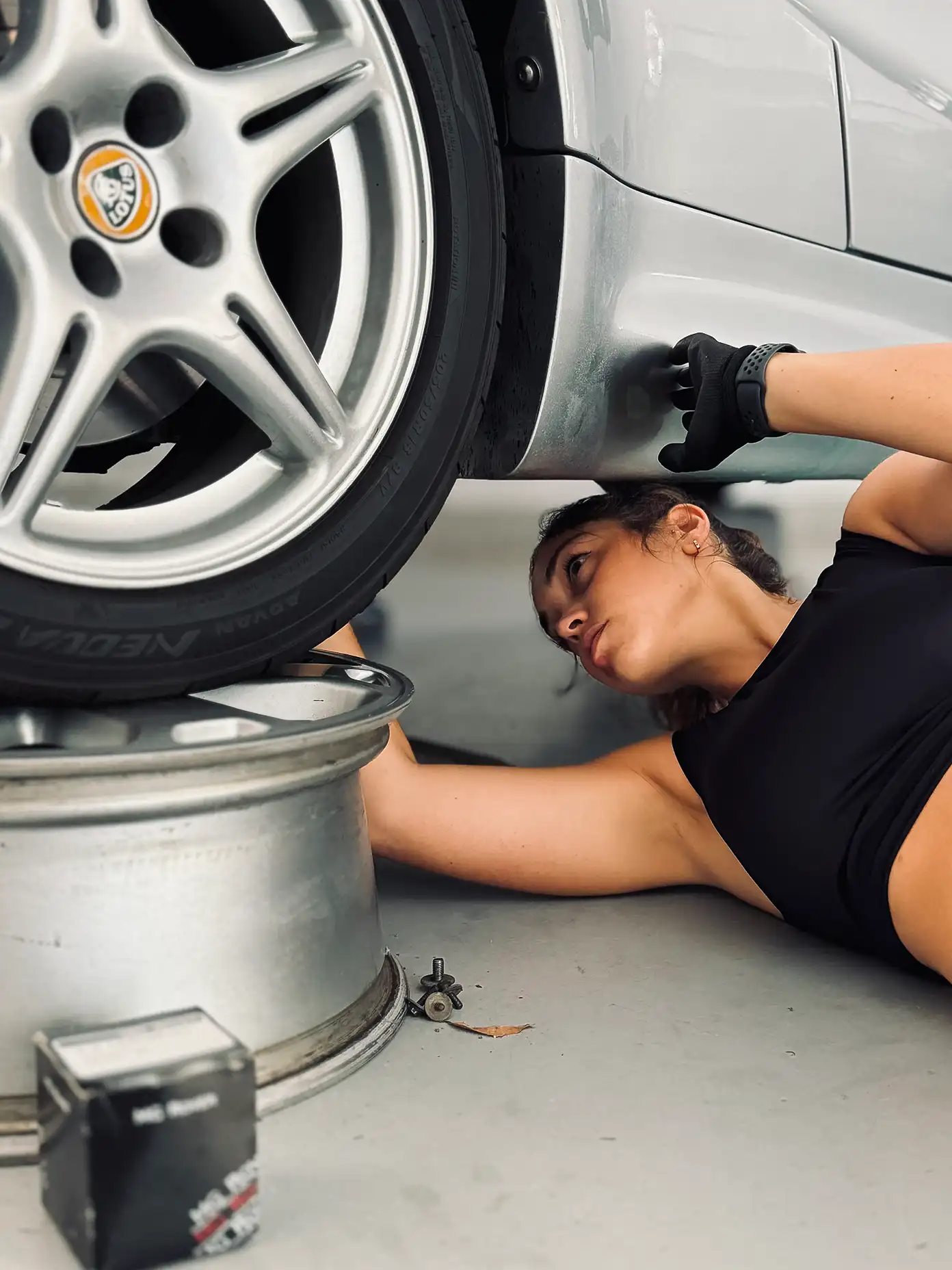 Elisa Artioli lying on the floor under a silver Lotus car, inspecting the underside near the wheel, with tools and a box nearby.