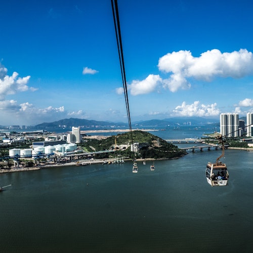 Kötélpályás felvonók lebegnek egy víztömeg felett, háttérben épületek, dombok és egy híd látható tiszta kék ég alatt.