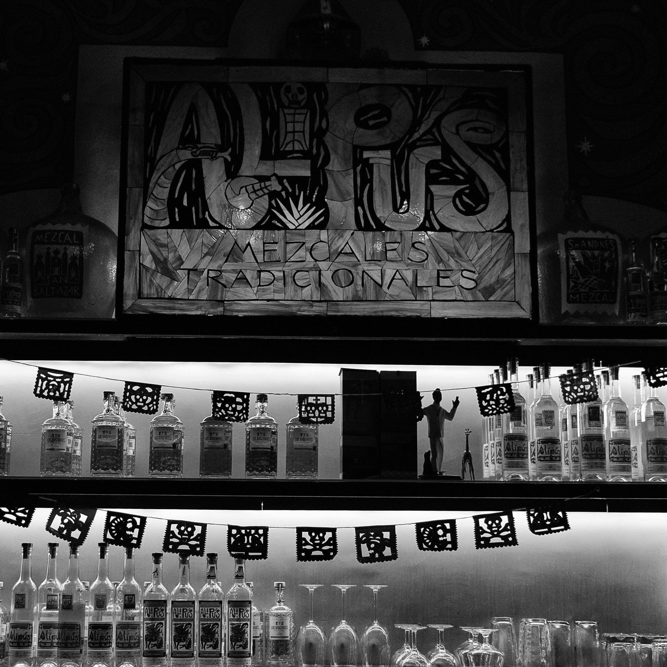 Black and white photo of a mezcal bar with backlit bottles, hanging papel picado, and a hand-painted sign reading “Alipús Mezcales Tradicionales.”