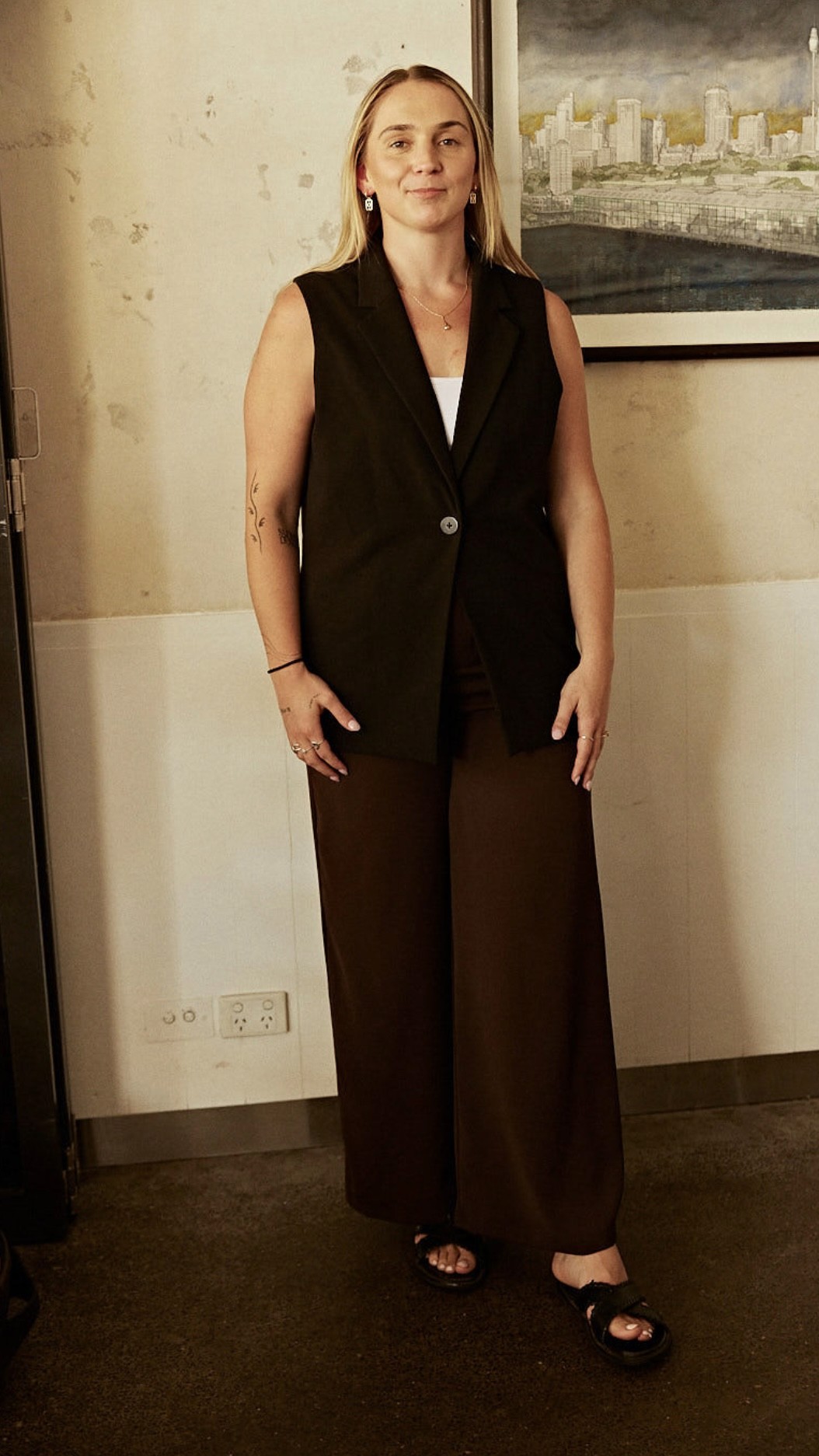 Person wearing black clothing carrying a black tote bag with a green globe-like geometric symbol, standing against a concrete wall in sunlight and shadow.