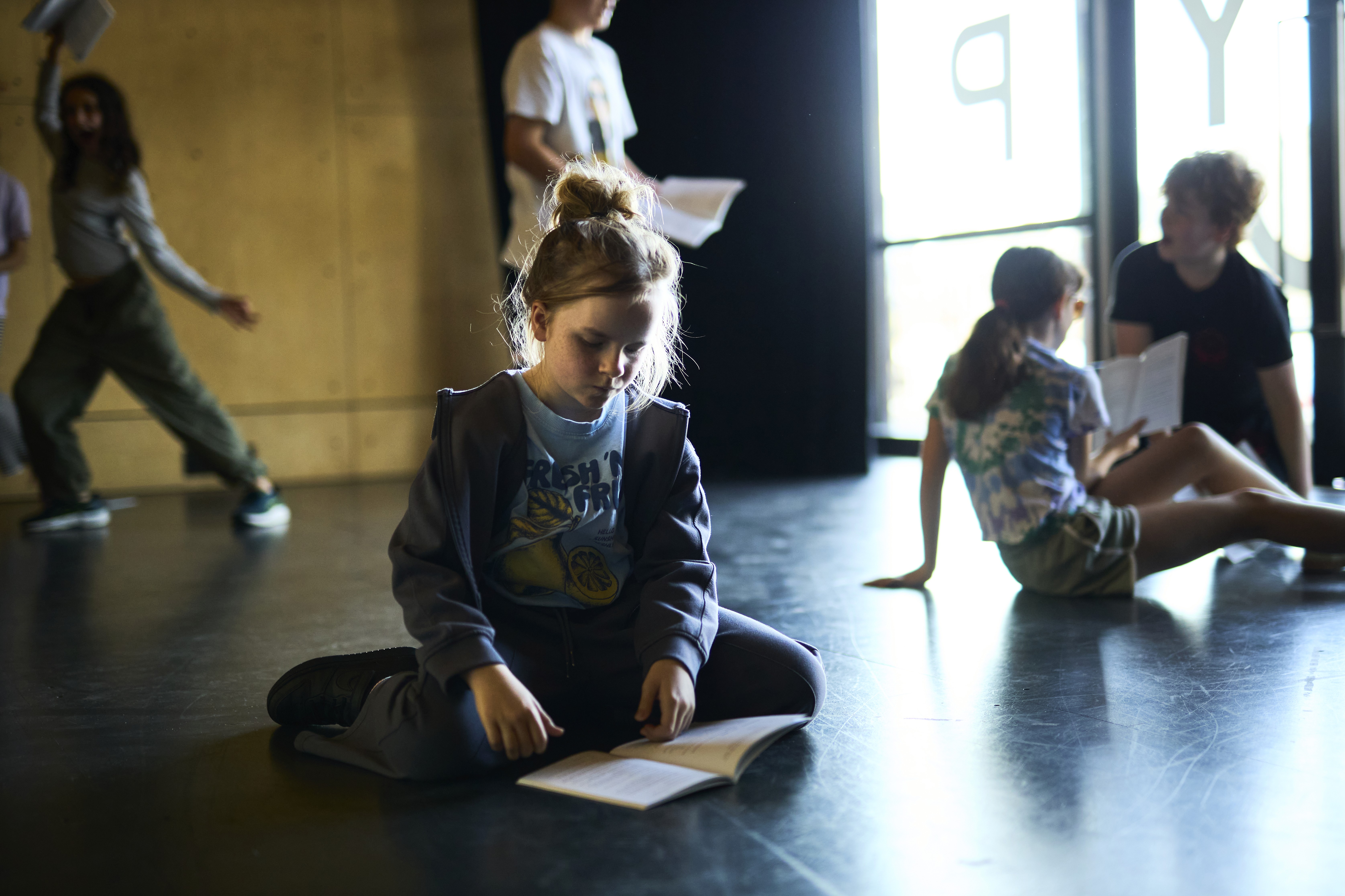 A girl sits cross legged on the floor reading a play.