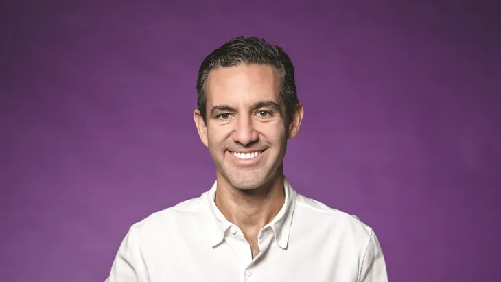 Smiling person with curly hair, wearing a black shirt, against a warm, dark background.