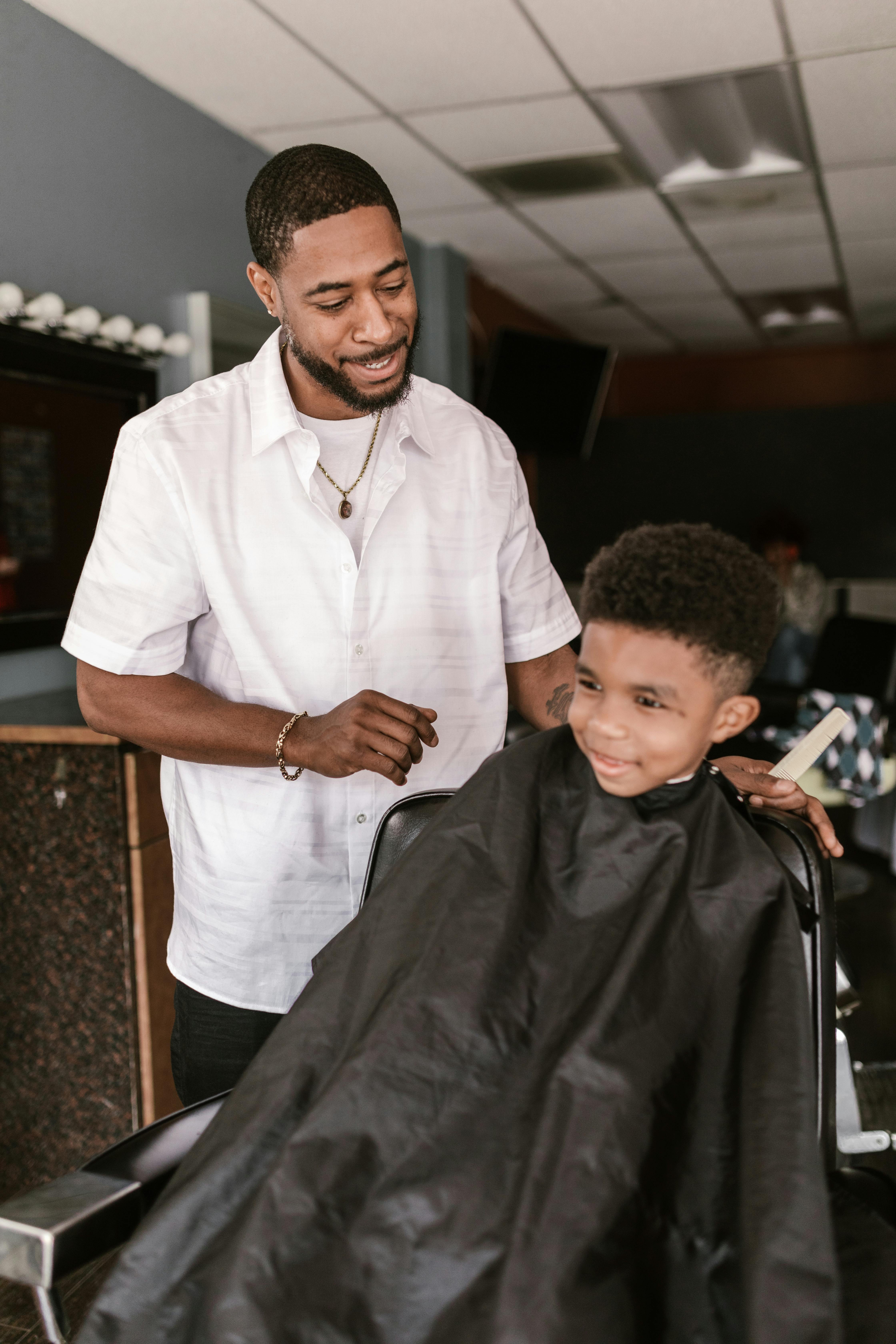 Barber with a young client getting a haircut. The barber, wearing a white shirt, smiles at the child.