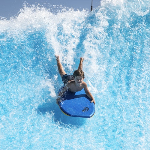 A person rides a bodyboard down a wave in a water park, with blue water splashing around them.