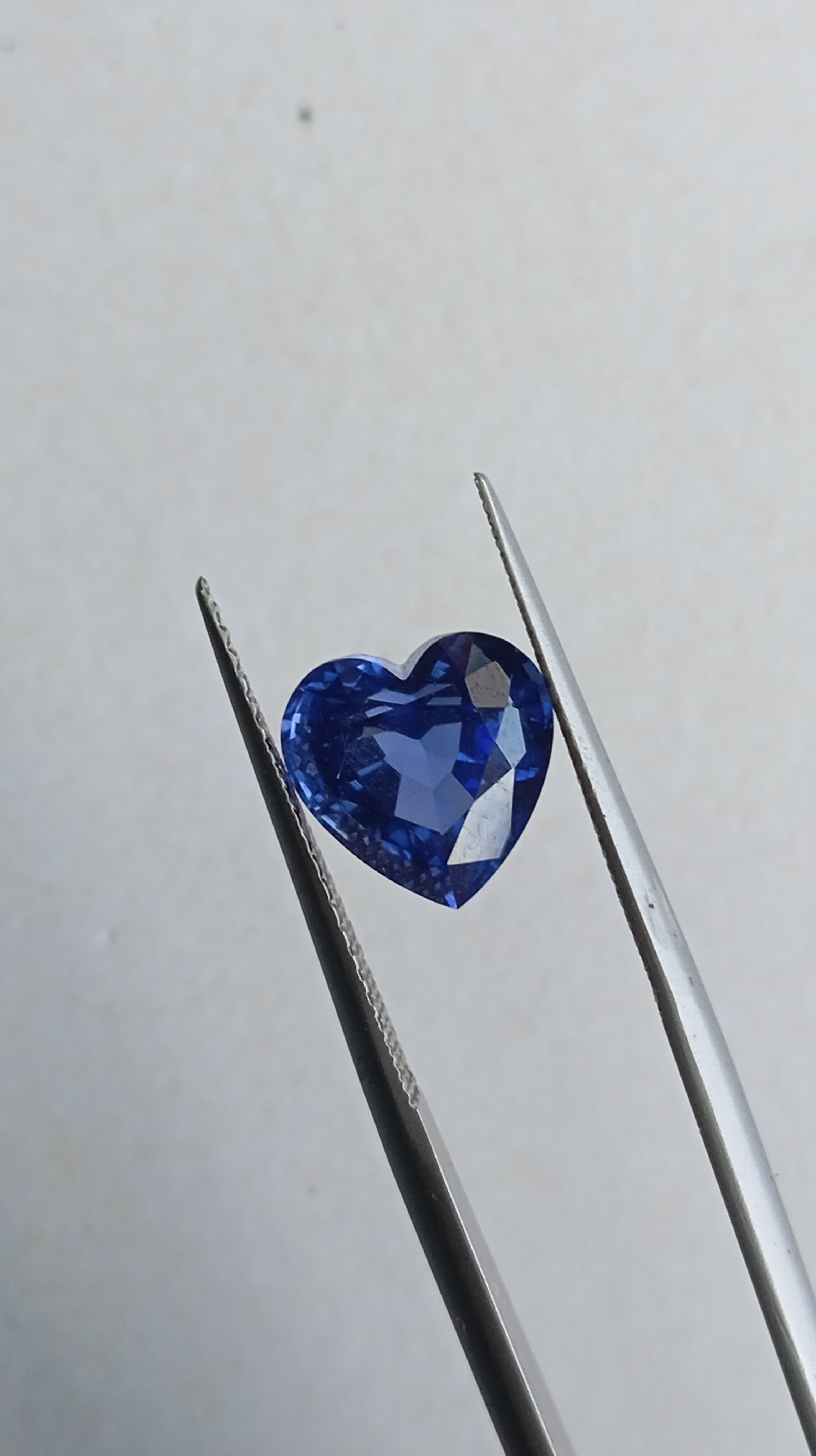 A vivid royal blue heart-shaped lab-grown sapphire gemstone held in tweezers for scale.