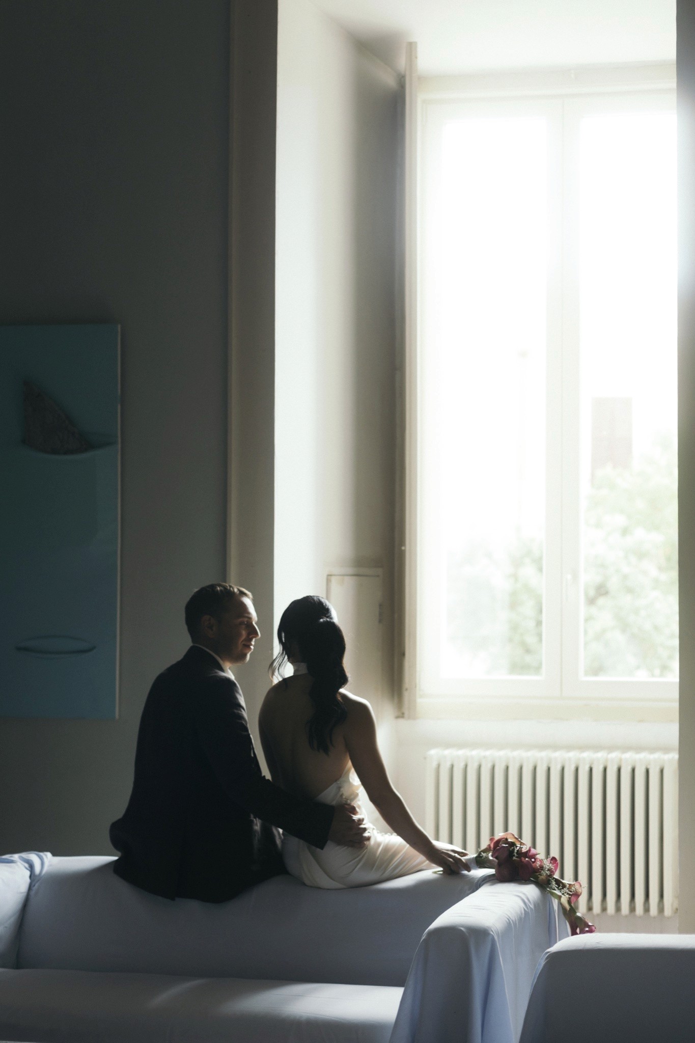 Toey and Otto seated together on a sofa arm beside a tall sunlit window