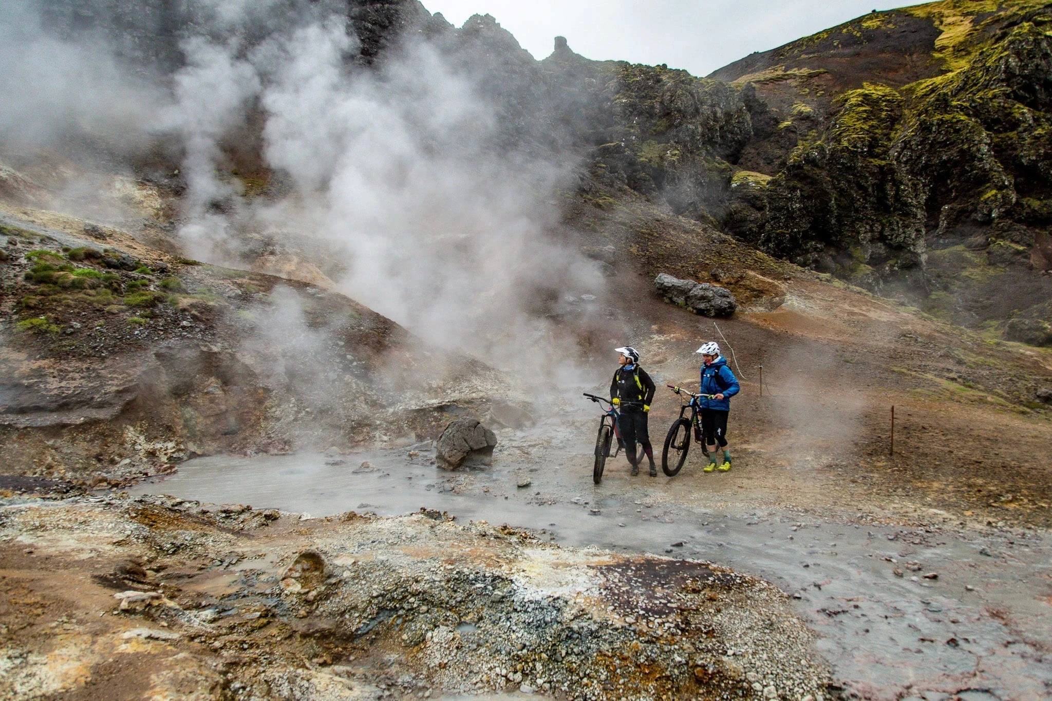 Two cyclists standing with bikes beside a steaming geothermal area.