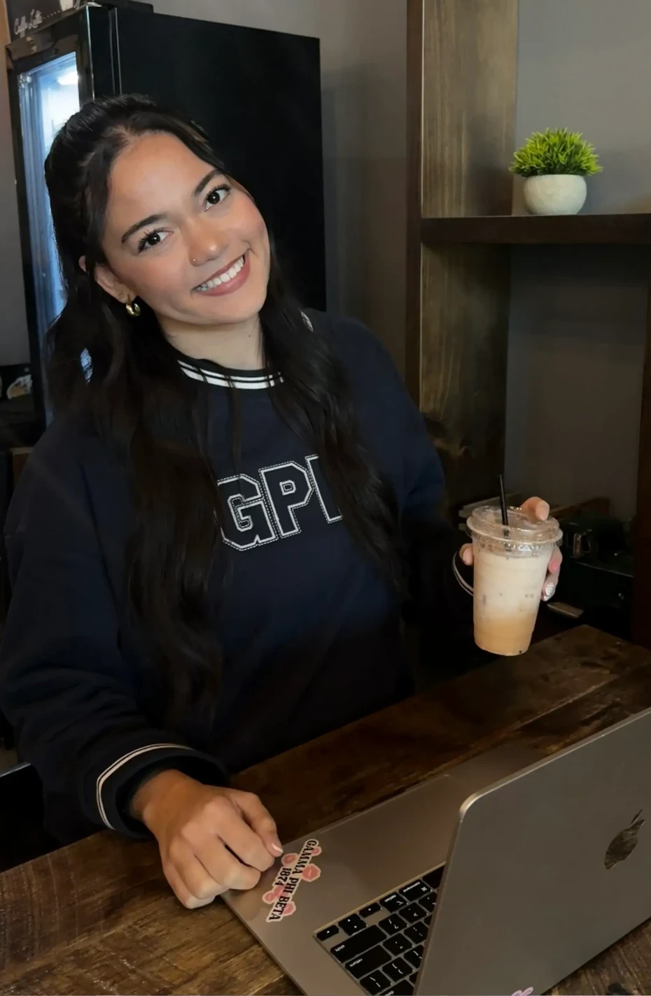 Person smiling at a café counter with a laptop open, holding an iced drink; wearing a dark “GPHI” crewneck