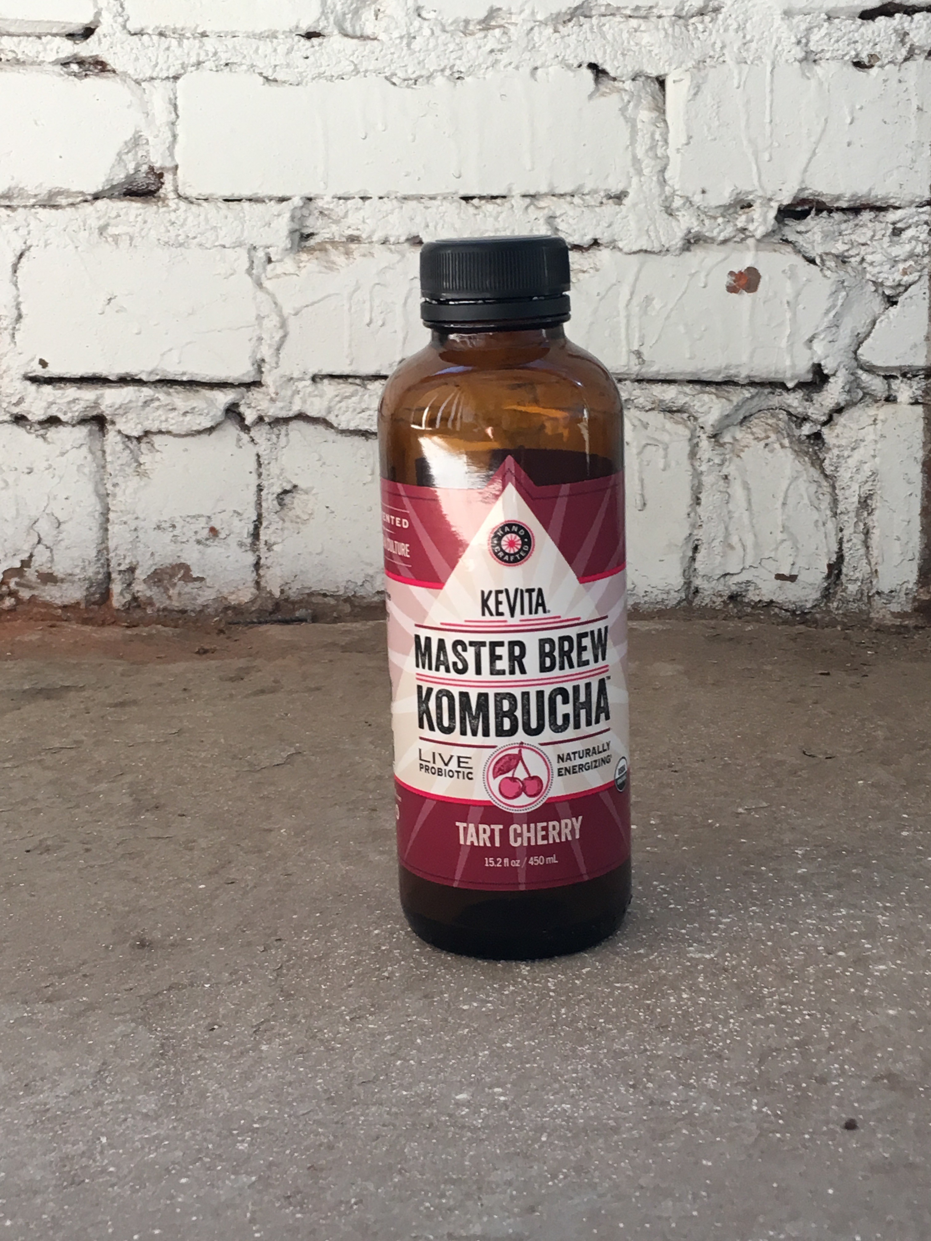 A brown bottle of Kevita Master Brew Kombucha Tart Cherry flavor is placed against a textured white brick wall on a concrete floor, showcasing its vibrant label design.
