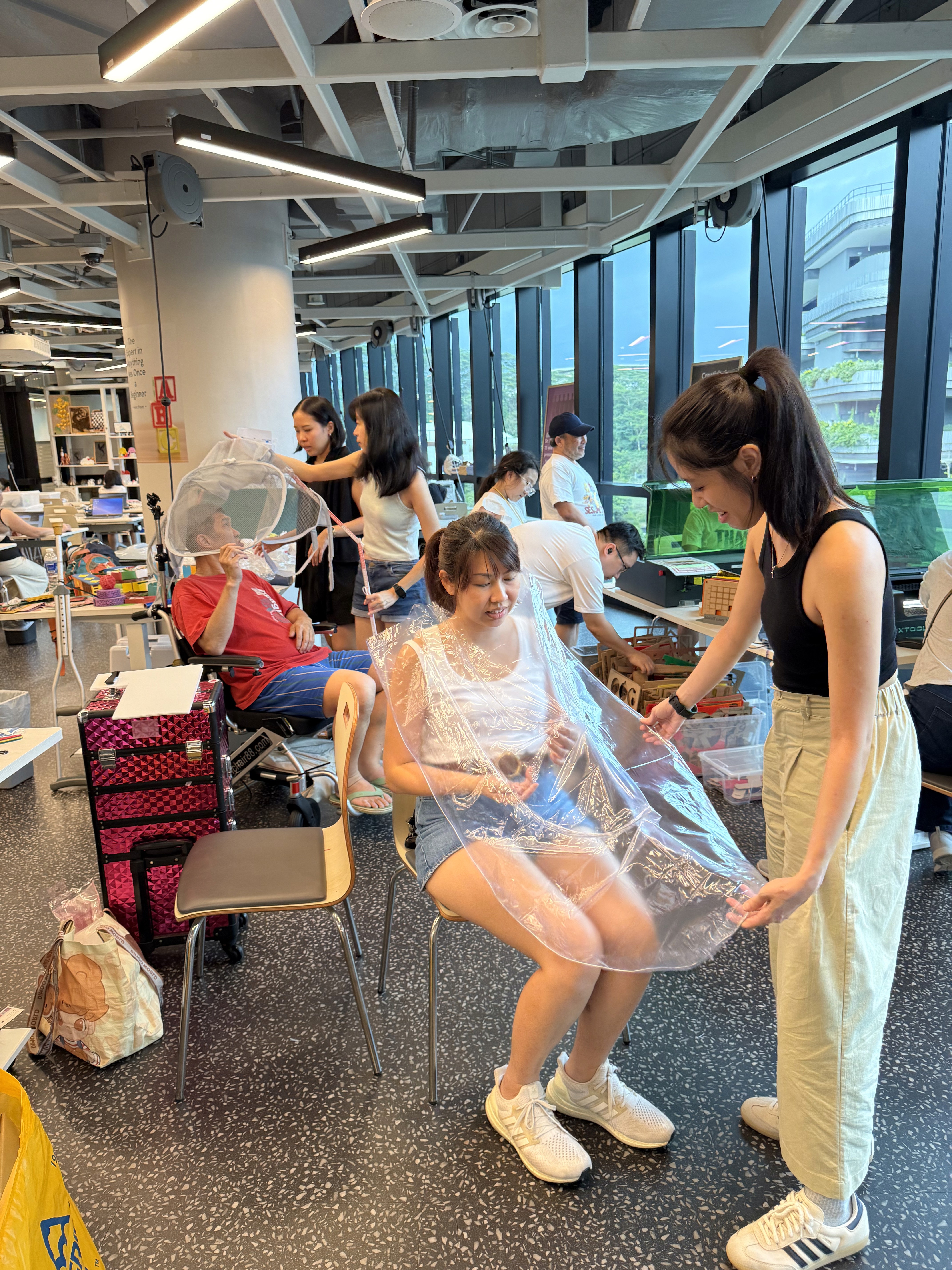 A woman sitting on a chair with a plastic cover over her and another woman draping it as they discuss designing rain cover for those in wheelchairs