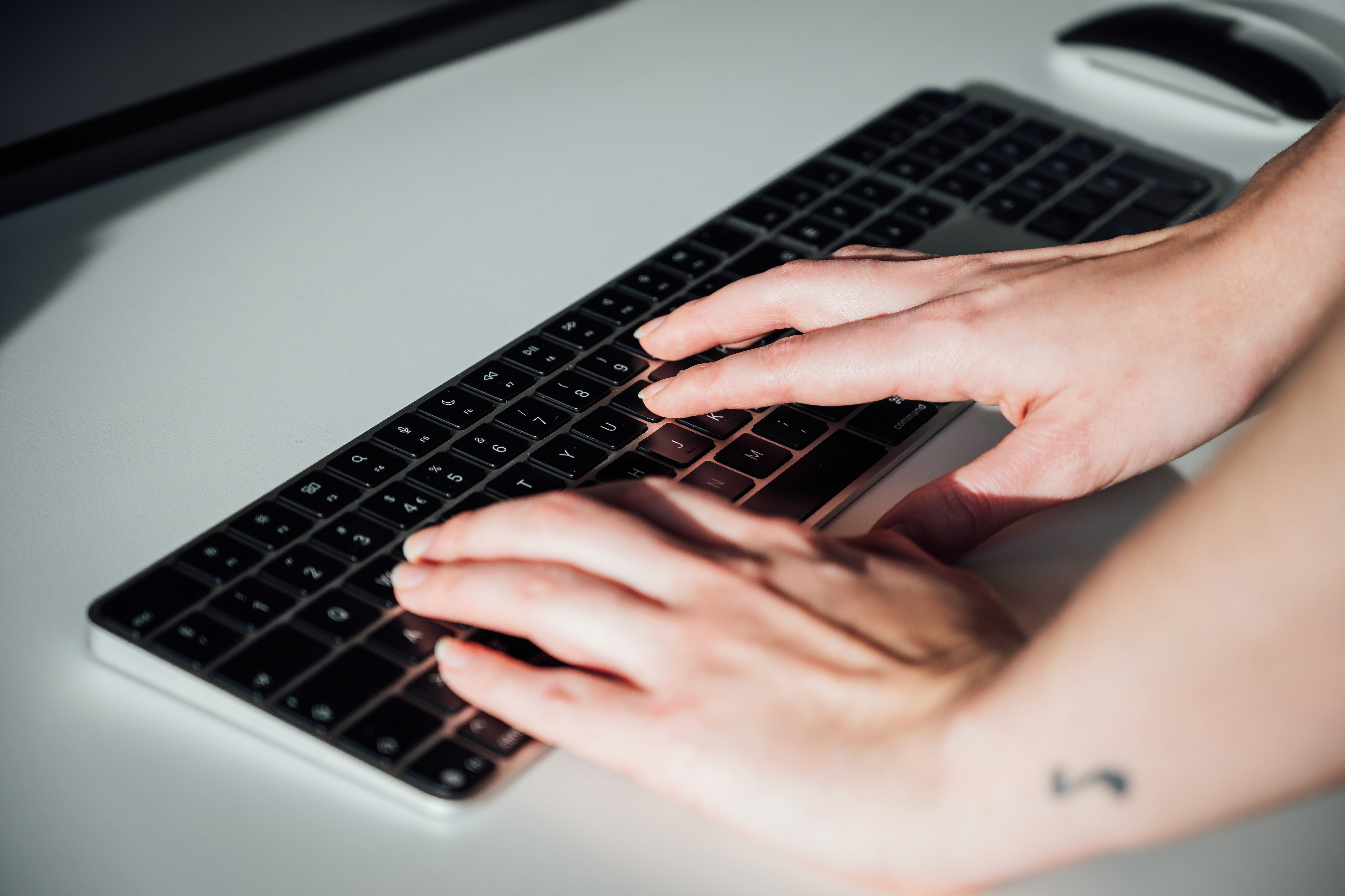 Nærbilde av hender som skriver på et svart tastatur på en lys bordflate, med en datamus i bakgrunnen.