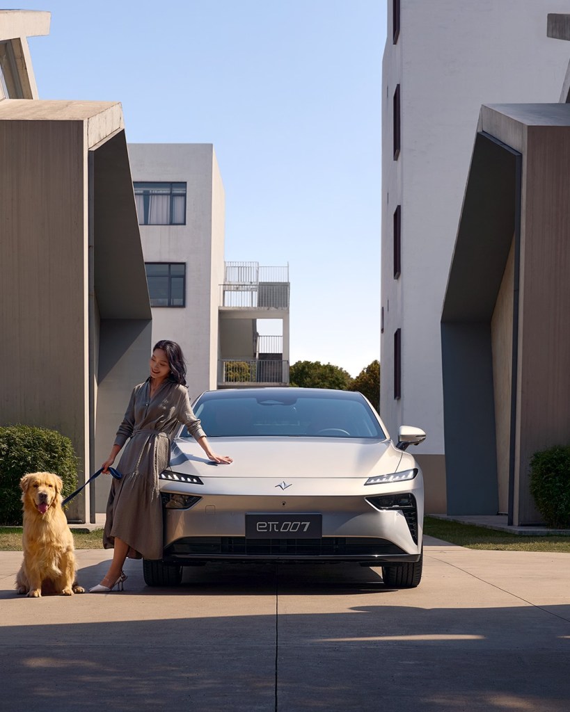 A woman in a modern dress stands next to a sleek, silver electric car while holding a leash of a golden retriever. The scene is set between two contemporary buildings under a clear blue sky.