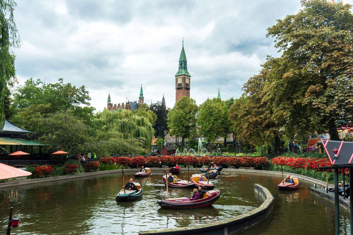 People in colorful swan-shaped paddle boats on a pond in front of a historic castle with green spires and gardens.