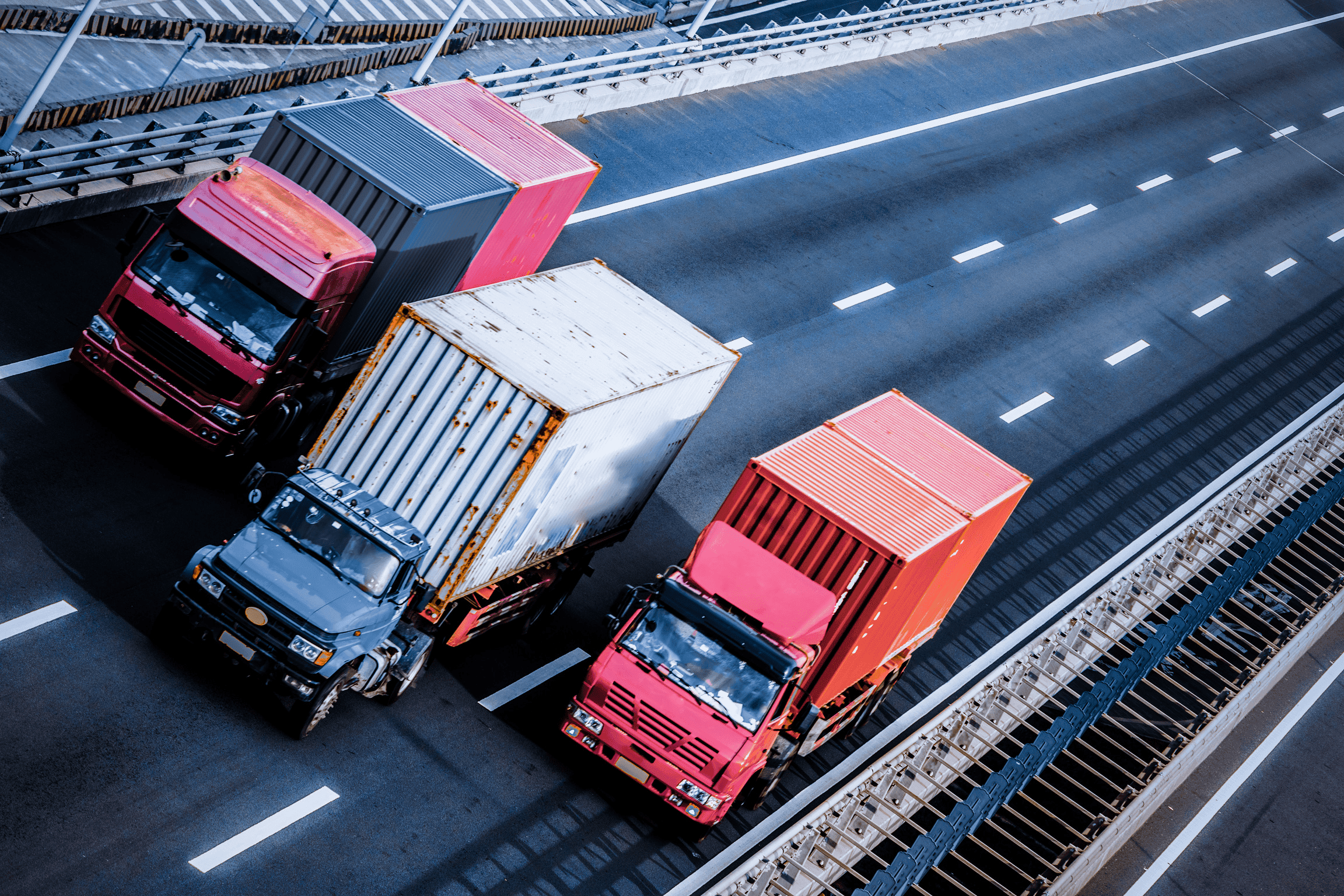 photo of three trucks driving on a highway expressway, side by side (each truck in a lane). The trucks on the ends are red and the truck in the center has a blue cabin with a white cargo area