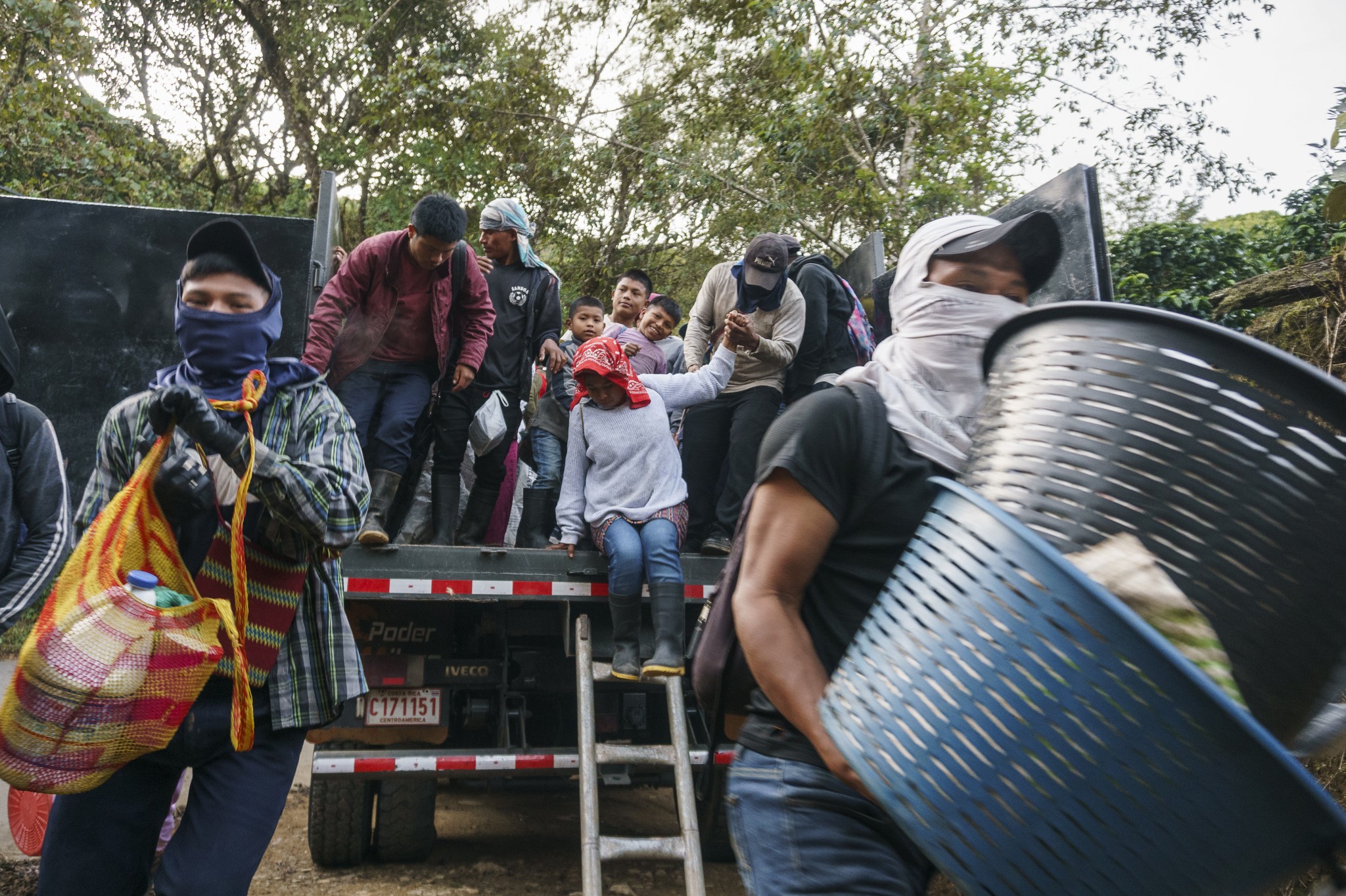 Workers, wearing hats and boots, unload from the back of a blue truck in a rural setting with trees and clear skies. One person is jumping down, holding a large machete. Others gather around or adjust their gear.