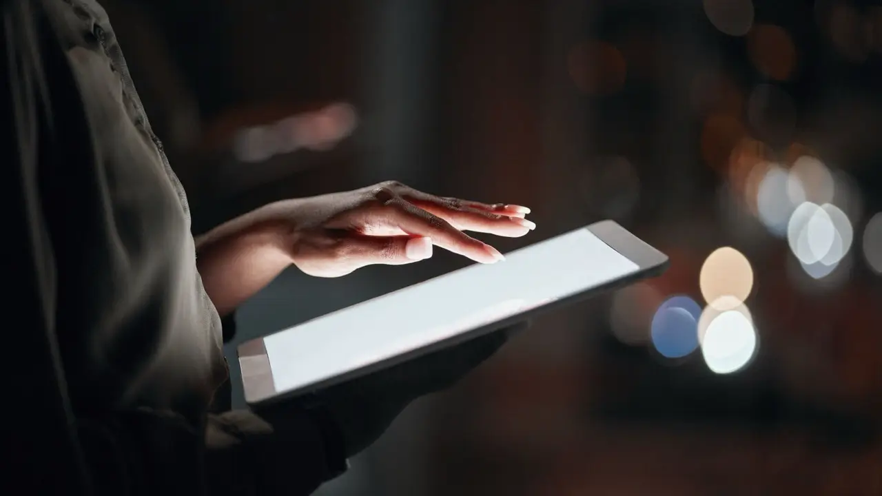 A person touches a glowing tablet screen in a dimly lit environment, with blurred lights in the background creating a bokeh effect, highlighting technology and digital interaction.