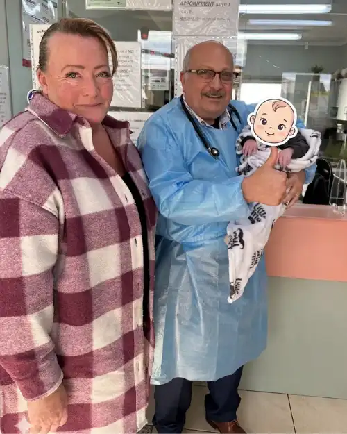 Dr. Ashraf Saddik giving a thumbs-up while cradling a wrapped infant next to a smiling family member near the reception desk in Coburg.