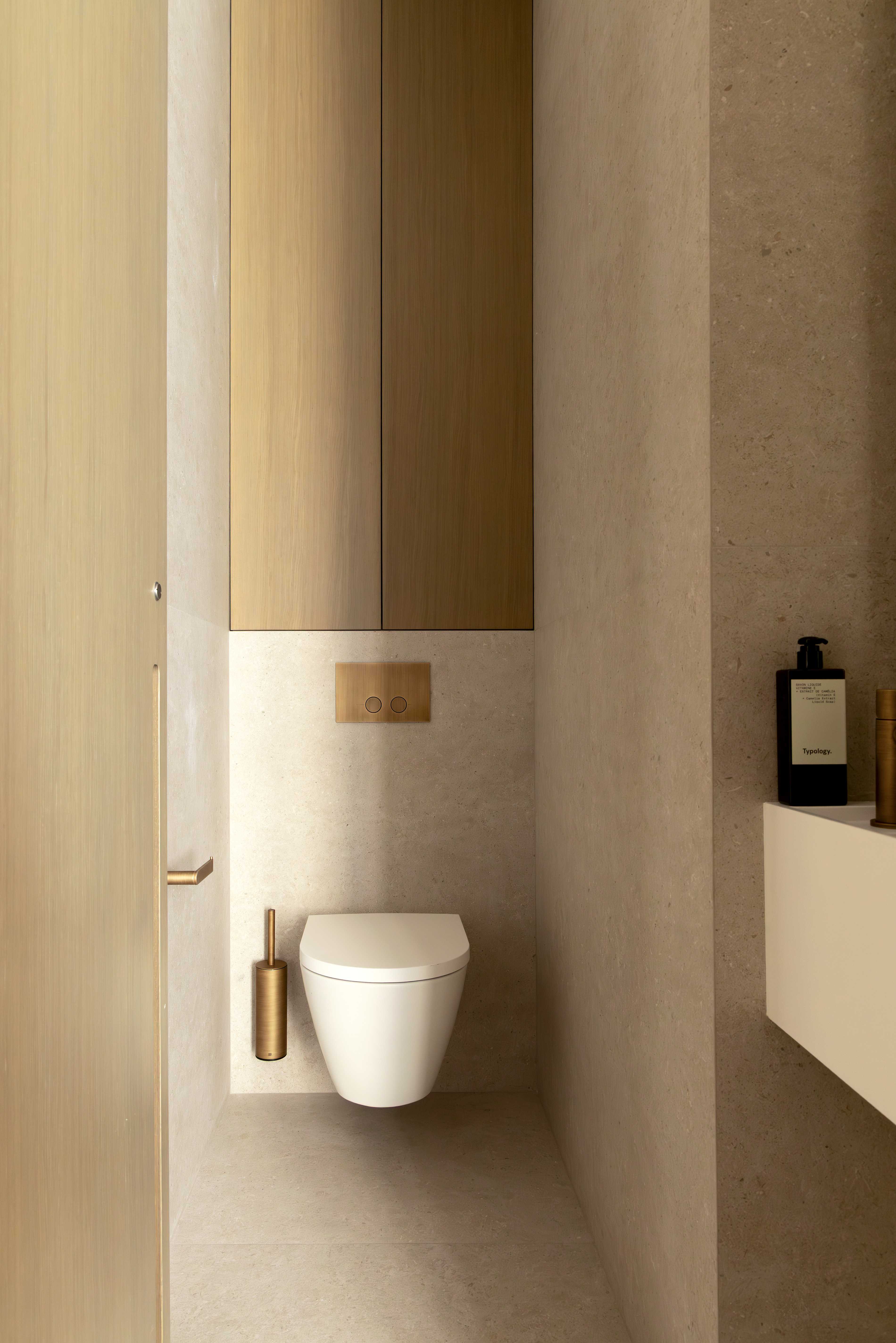 Detail WC suspendu avec plaque doree, rangement haut en bois, murs en pierre et produits sur etagere laterale