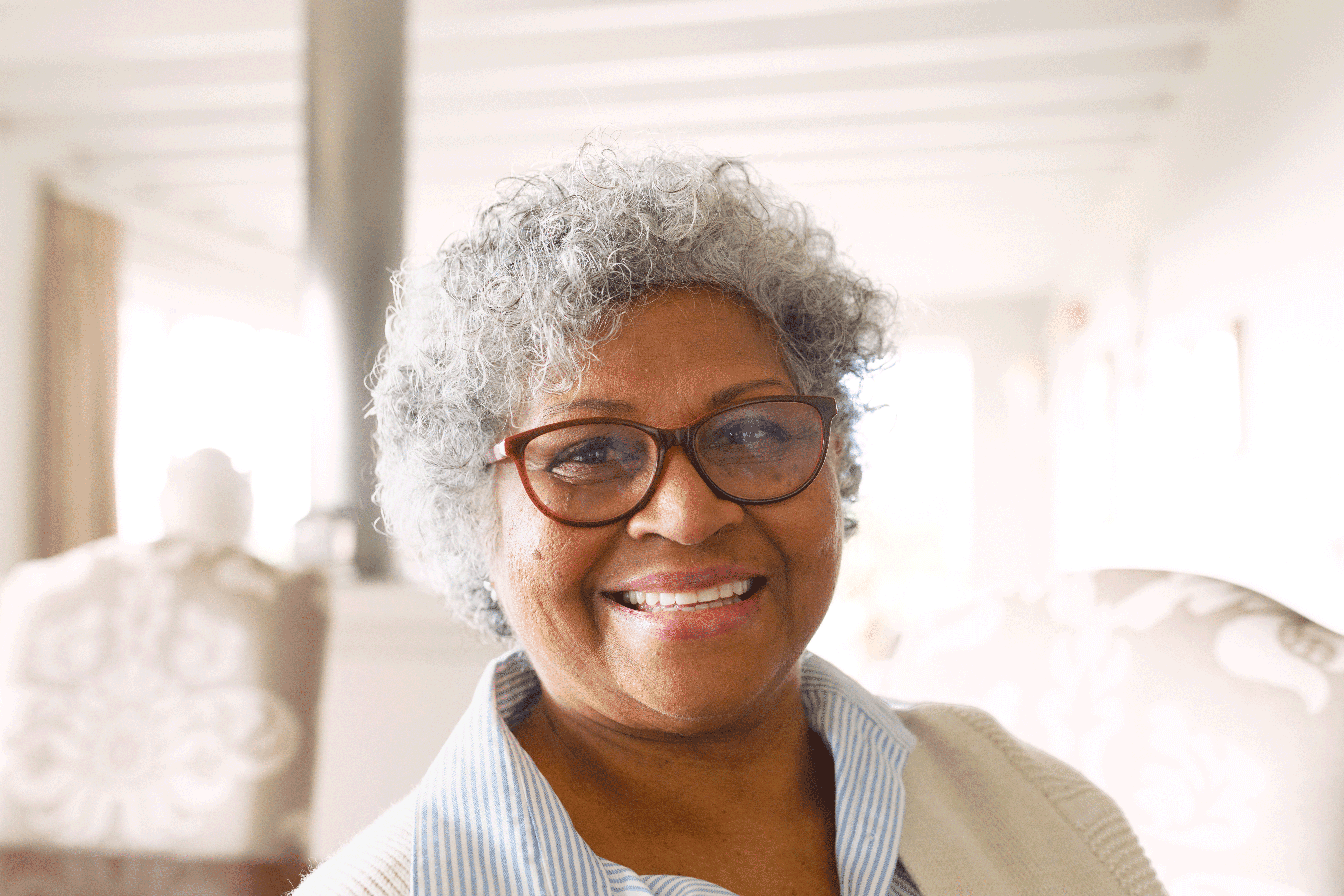 A smiling woman with curly grey hair and glasses looks directly at the camera in a brightly lit indoor setting. She is wearing a blue and white striped shirt layered under a light-colored cardigan.