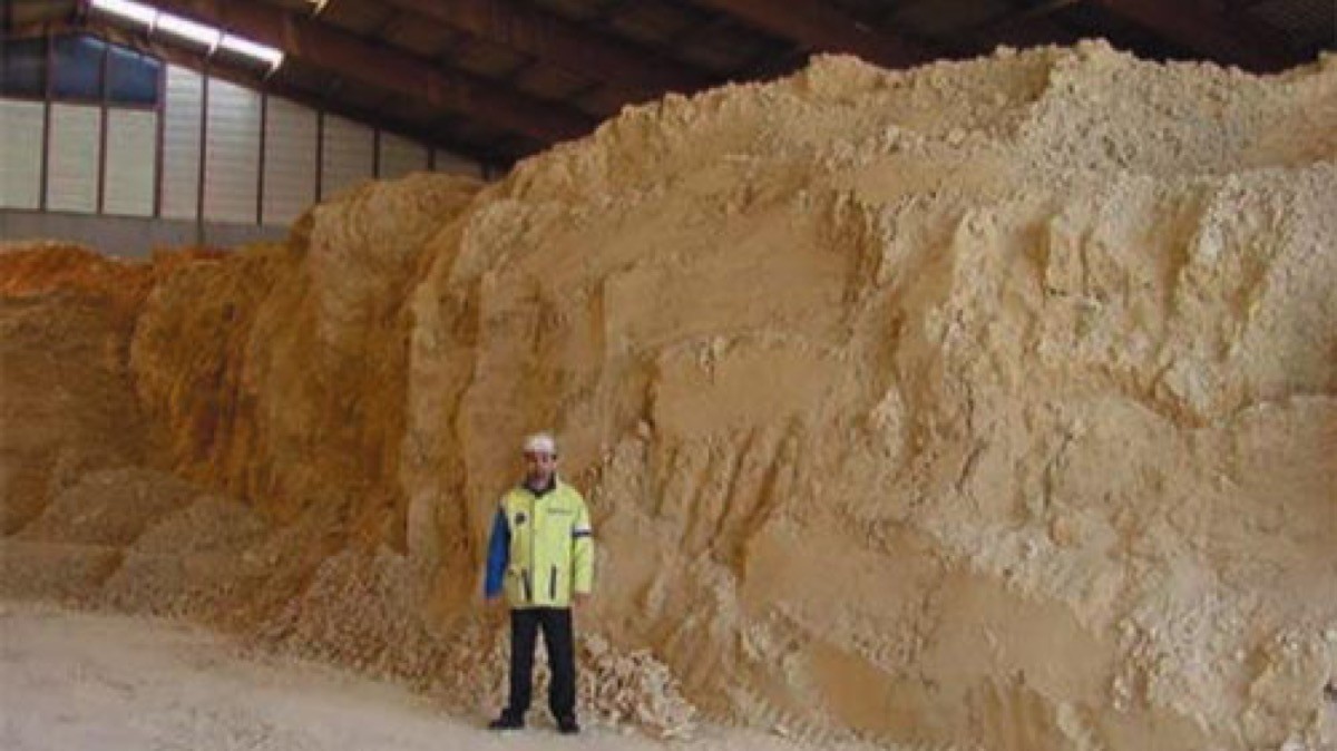 Radium-bearing waste at the La Rochelle plant: a man in a hard hat and a high-visibility jacket stands in front of a pile of pale yellow material that towers over him inside a warehouse