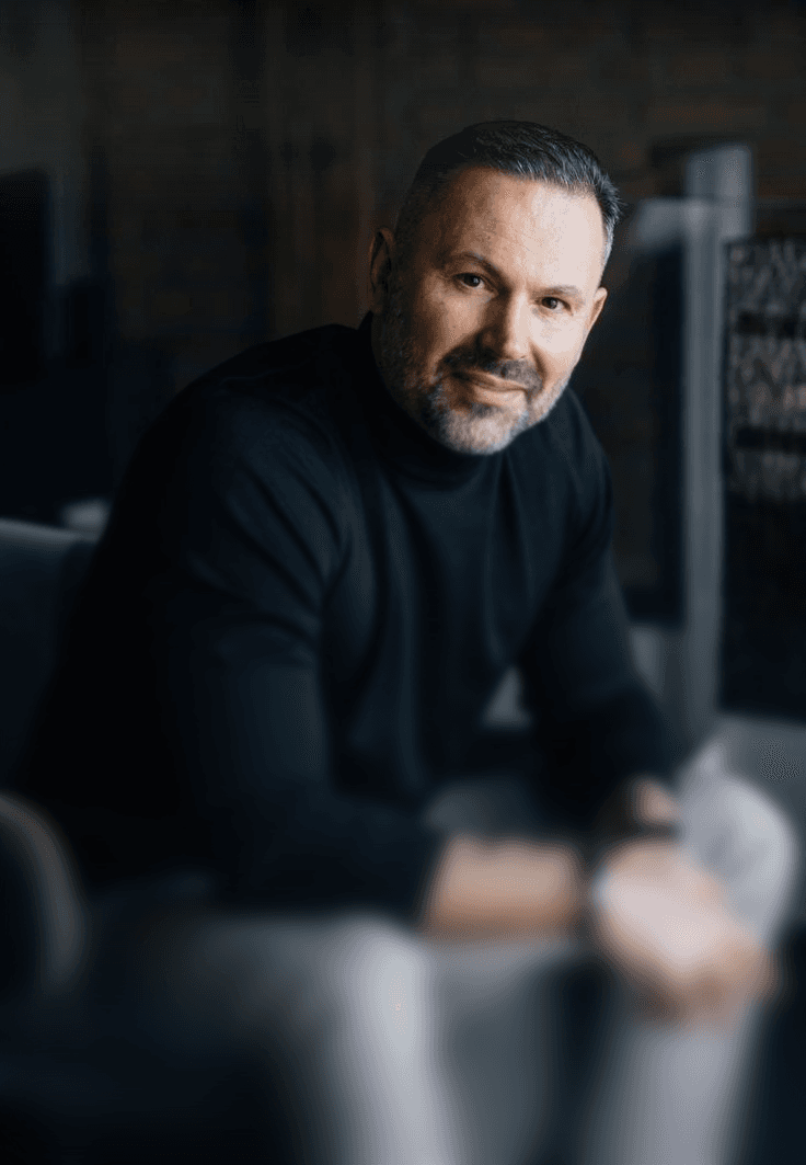 A man in a black turtleneck and gray pants sits casually, leaning forward with fingers intertwined. The mood is relaxed and contemplative.