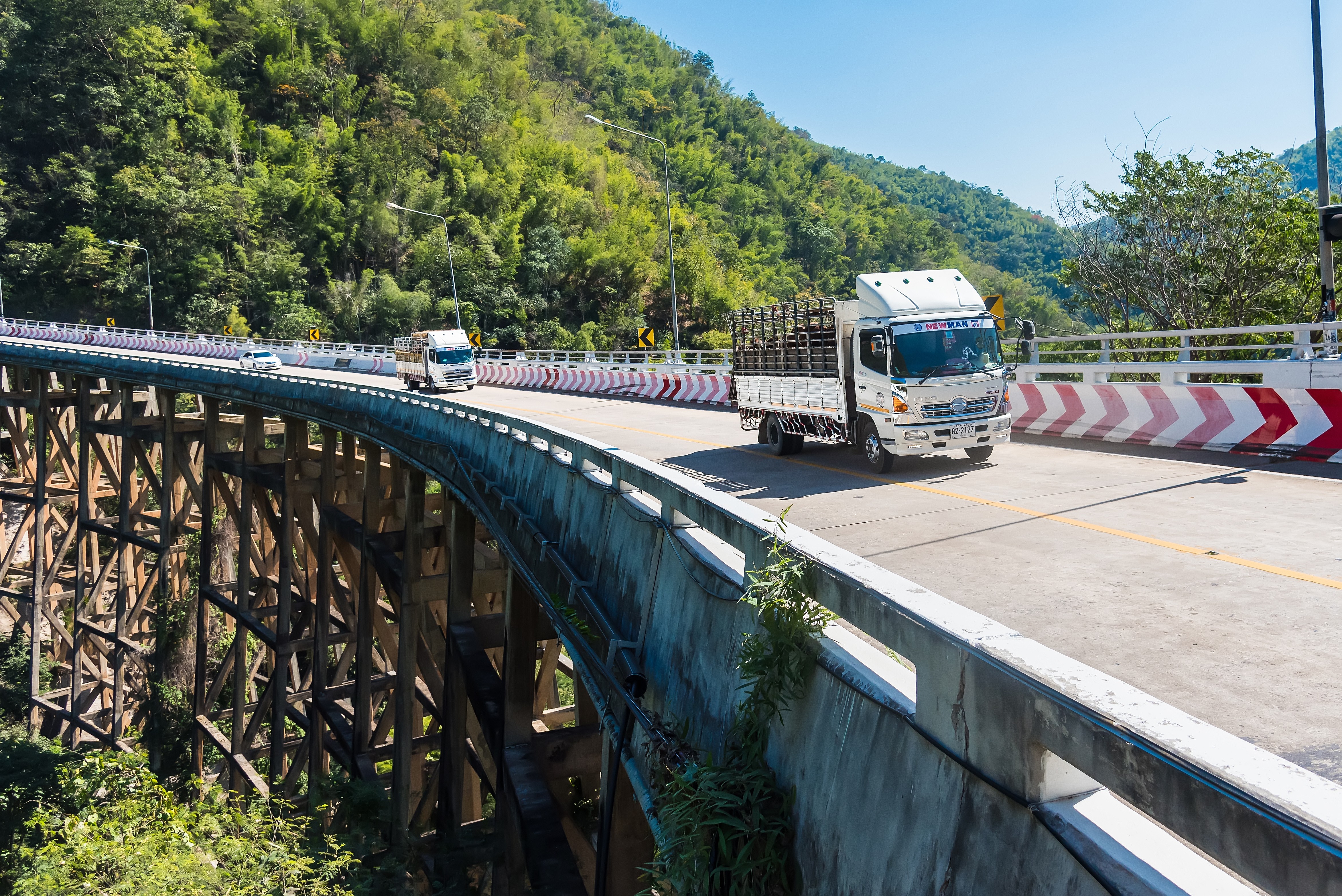 Cargo trucks driving across a curved bridge through a forested mountain landscape.