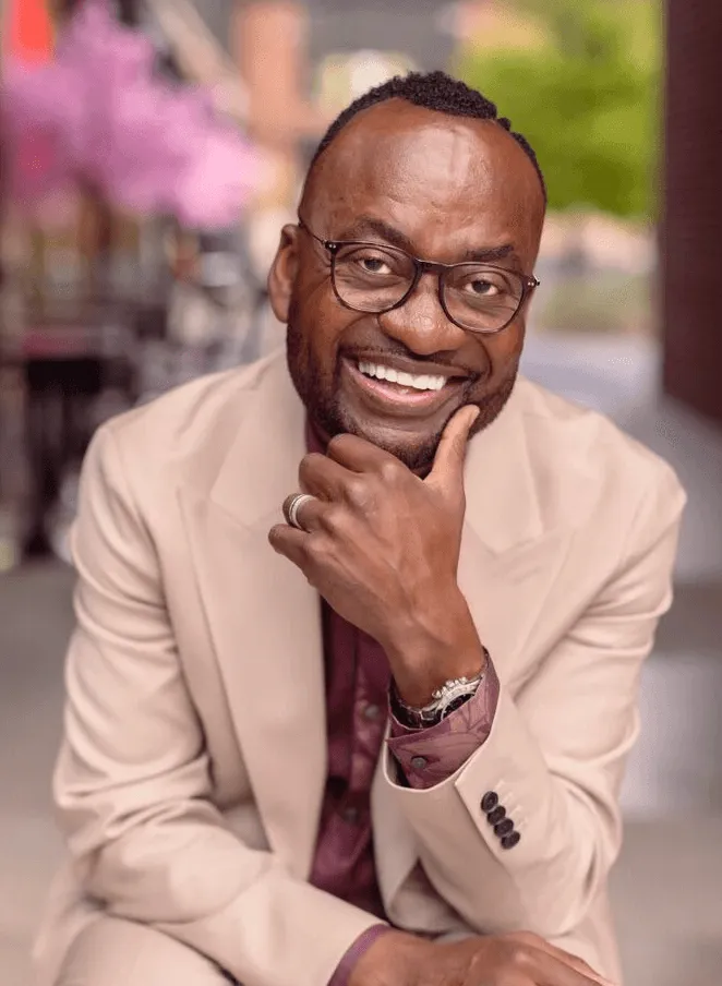 Smiling man in a beige blazer resting his chin on his hand.