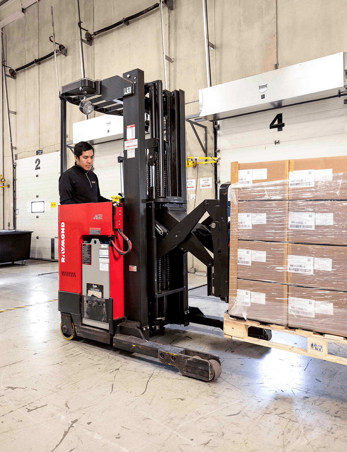 Warehouse worker operates a red forklift to move a wrapped pallet of boxes near loading bay doors.