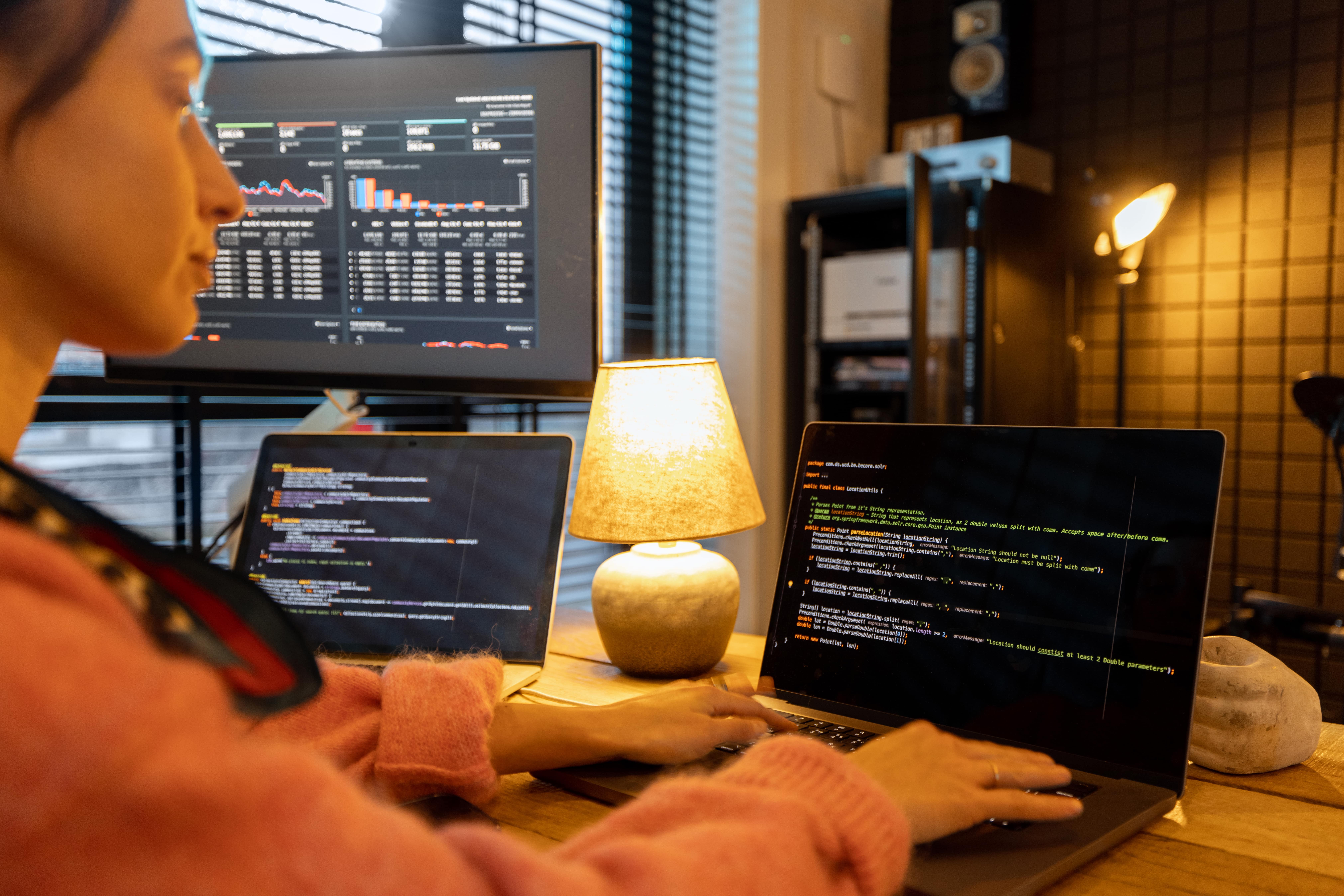 Female programmer working on multiple screens with code editor and data dashboard in a cozy, dimly-lit workspace.