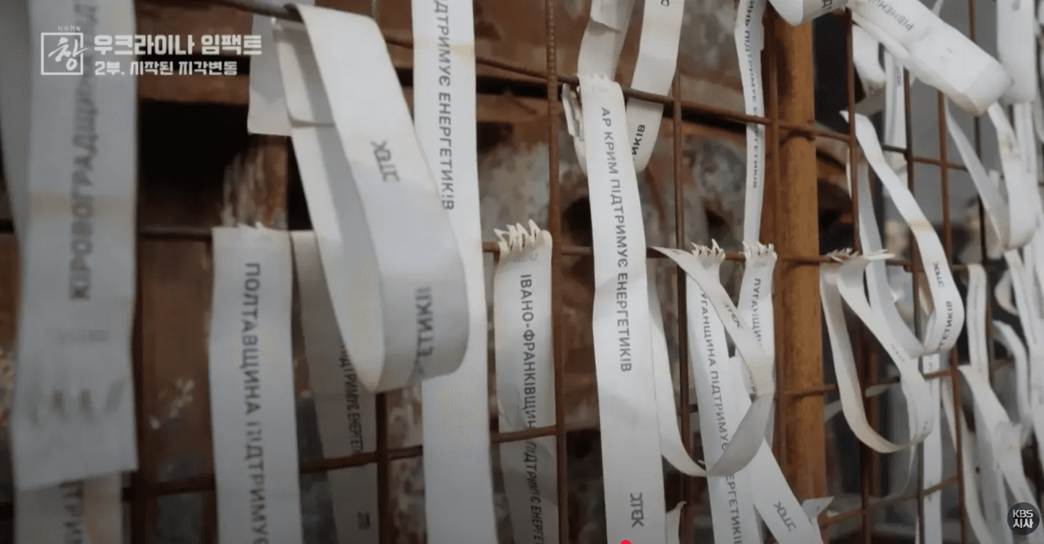 우크라이나 추모 리본들이 걸린 울타리. 전쟁 희생자들의 이름이 적힌 흰색 리본들이 여러 개 매달려 있음. KBS 시사기획 창 우크라이나 임팩트 2부 화면.