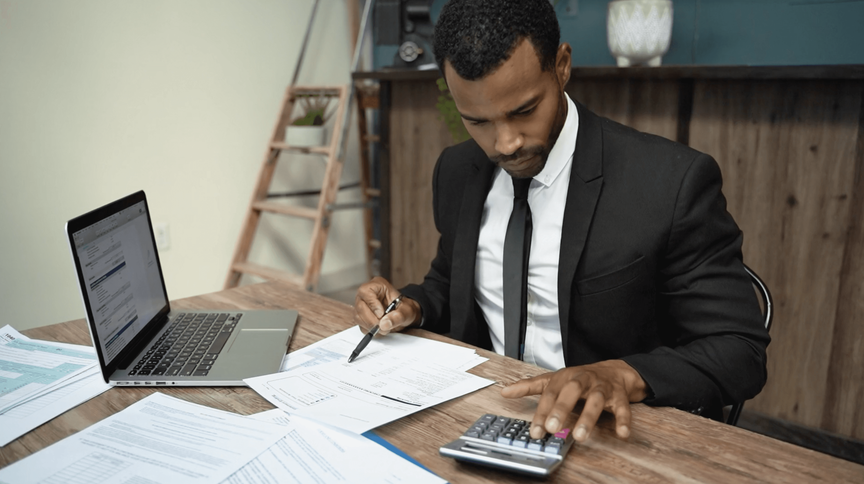 A man in a suit sits at a desk, analyzing documents and using a calculator next to a laptop.