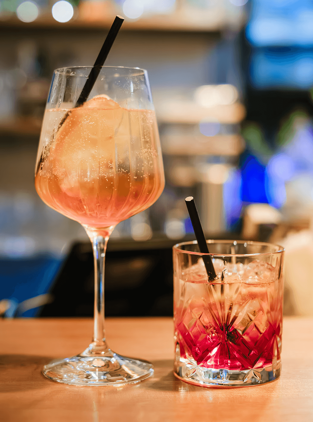 Two refreshing cocktails on a bar counter: a tall stemmed glass filled with a light orange spritz garnished with a large ice cube and a straw, beside a shorter glass containing a pink iced drink with a black straw, with a softly blurred bar background.