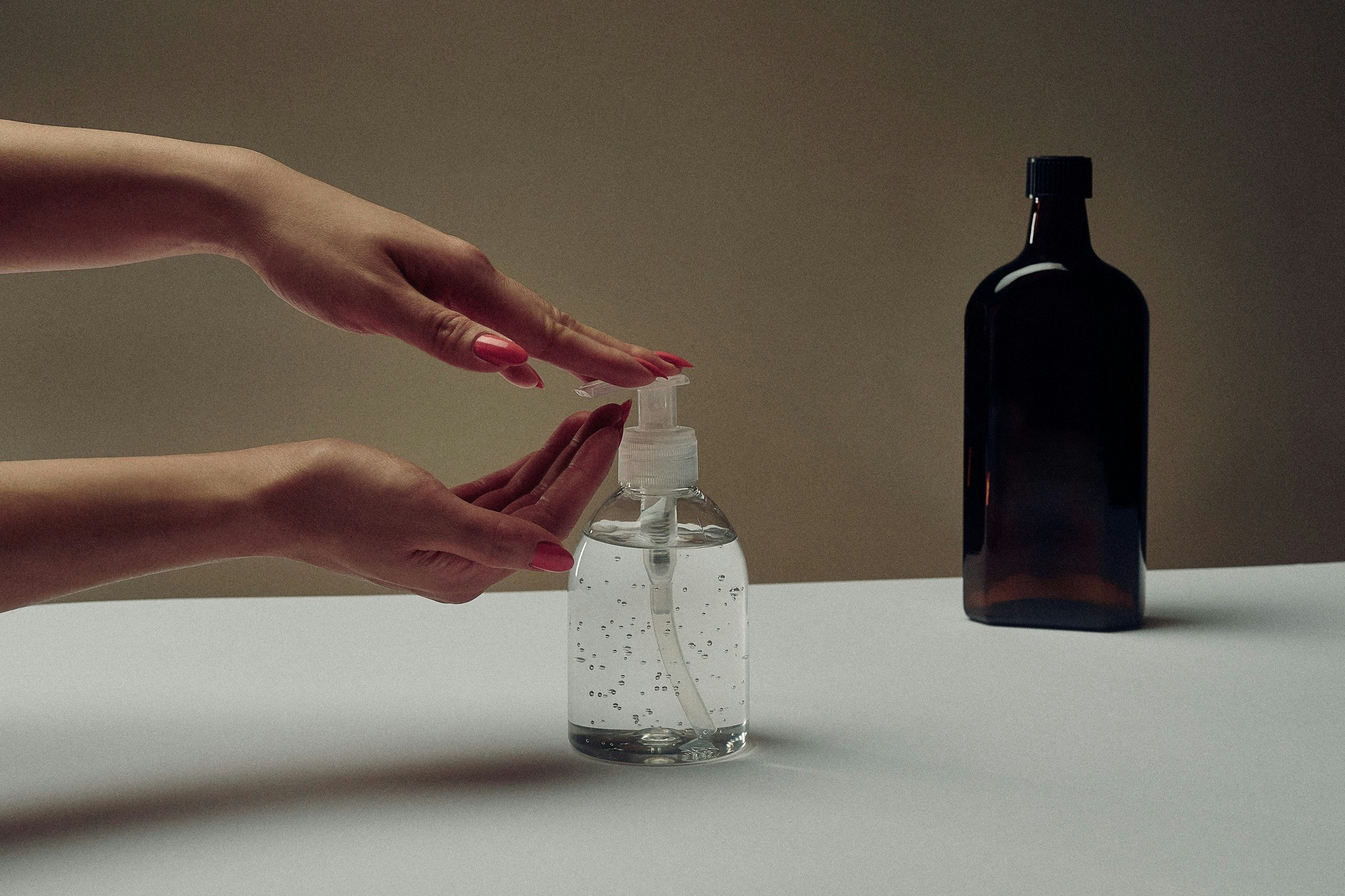Hands with painted nails use a clear hand sanitizer pump bottle on a white surface; a dark bottle stands in the background, evoking cleanliness.