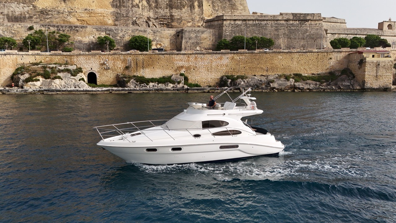 Sealine F37 motor yacht cruising through Grand Harbour Valletta Malta with historic limestone bastions in the background — white flybridge cruiser underway on blue Mediterranean water