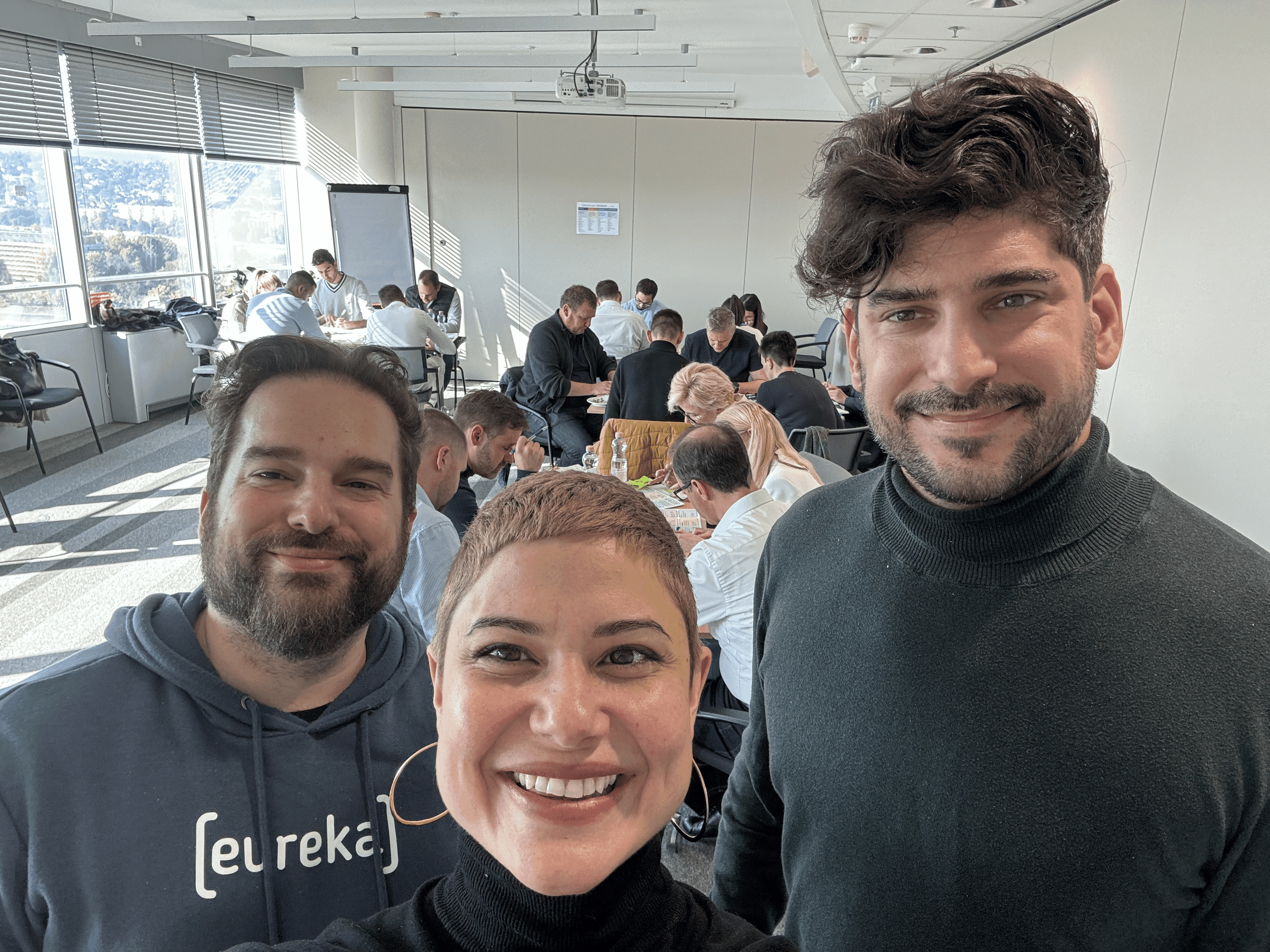 Three trainers smile in a room while a group solves tasks in the background.