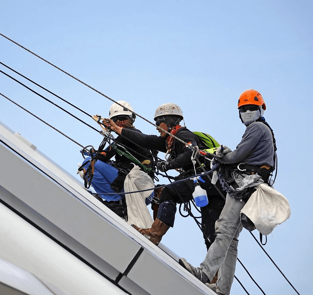 Three workers in safety gear and helmets are suspended by ropes, cleaning a modern building's slanted roof, against a clear blue sky.