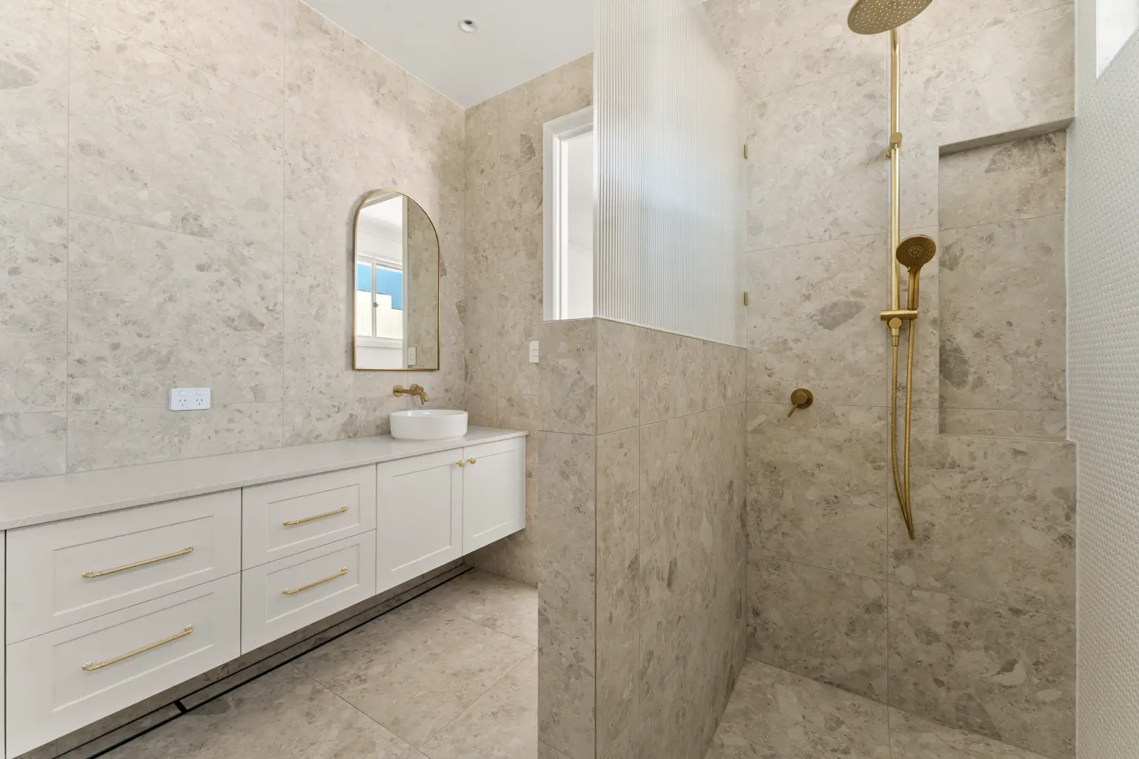 Modern luxury bathroom in a completed custom home featuring floor-to-ceiling stone-look tiles, a white vanity with brass hardware, an arched mirror, and a walk-in shower with a fluted glass screen.