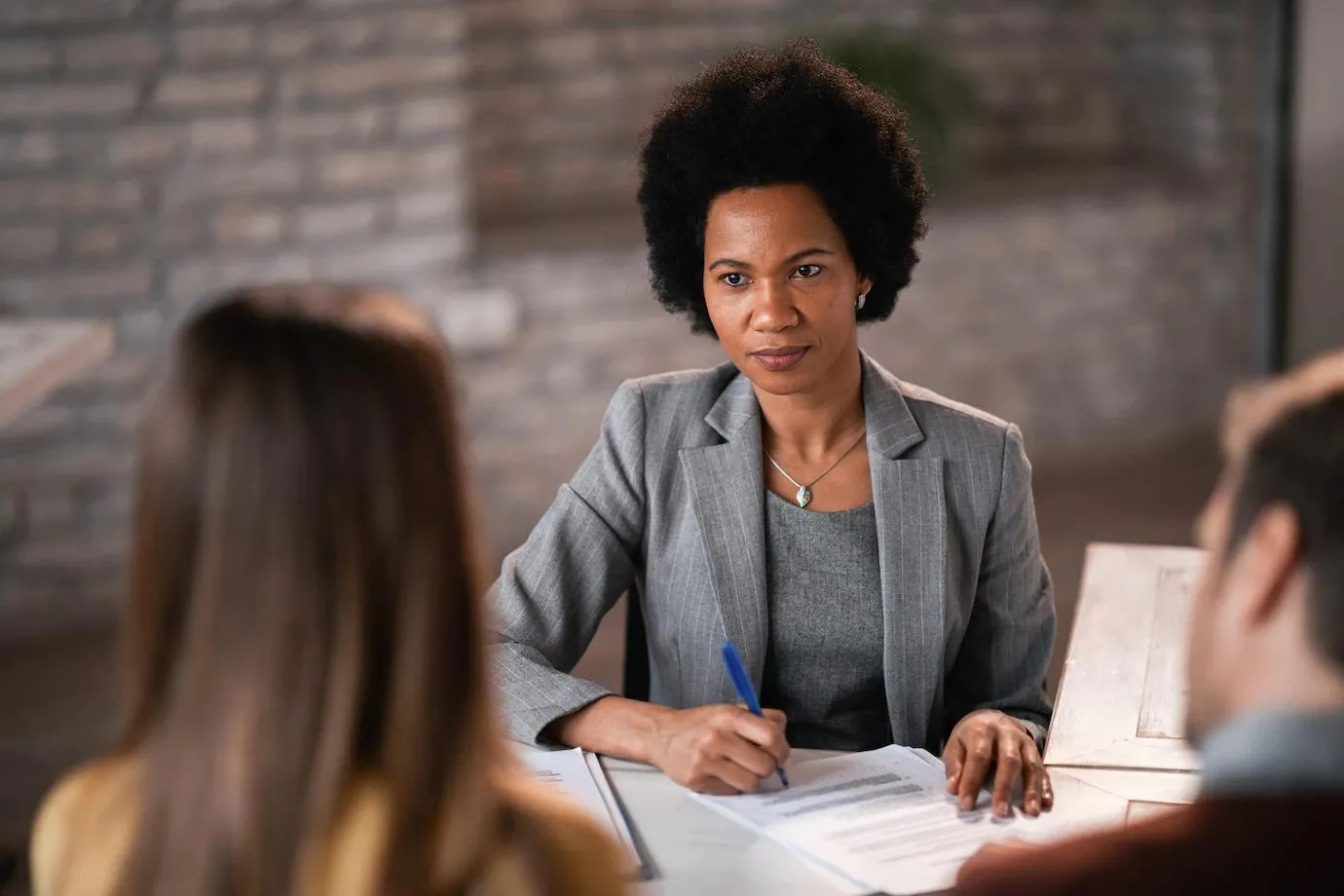 Mulher negra em ambiente profissional, vestida com um terno cinza, segurando uma caneta e analisando documentos enquanto conversa com duas pessoas, que aparecem de costas. A expressão séria da mulher e sua postura transmitem profissionalismo e atenção, representando uma situação de entrevista ou reunião de negócios.