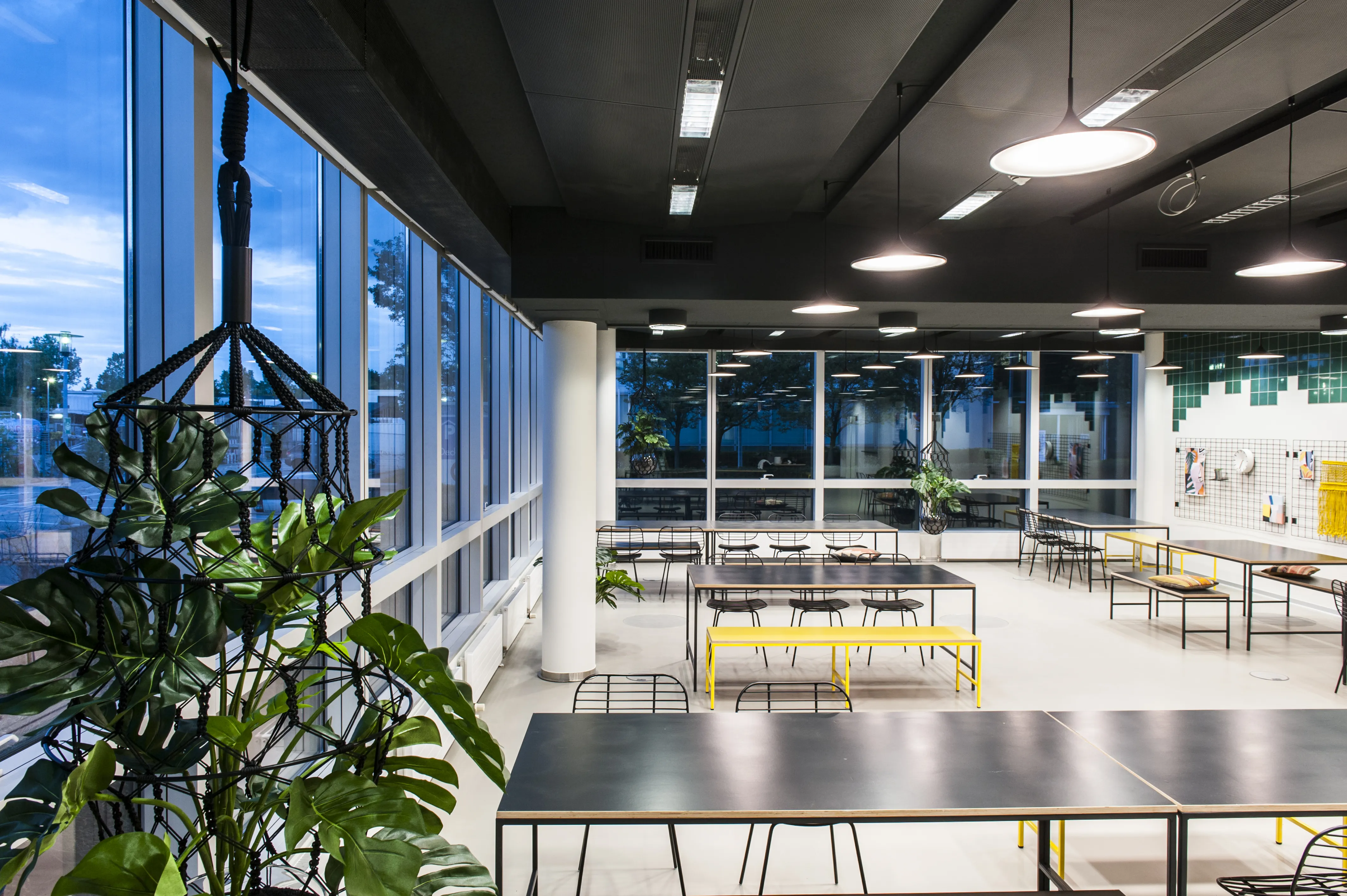 Panoramic evening view of KANTINE KOMTUR with floor-to-ceiling windows, black canteen tables, yellow benches, hanging plants and round white pendant lights.